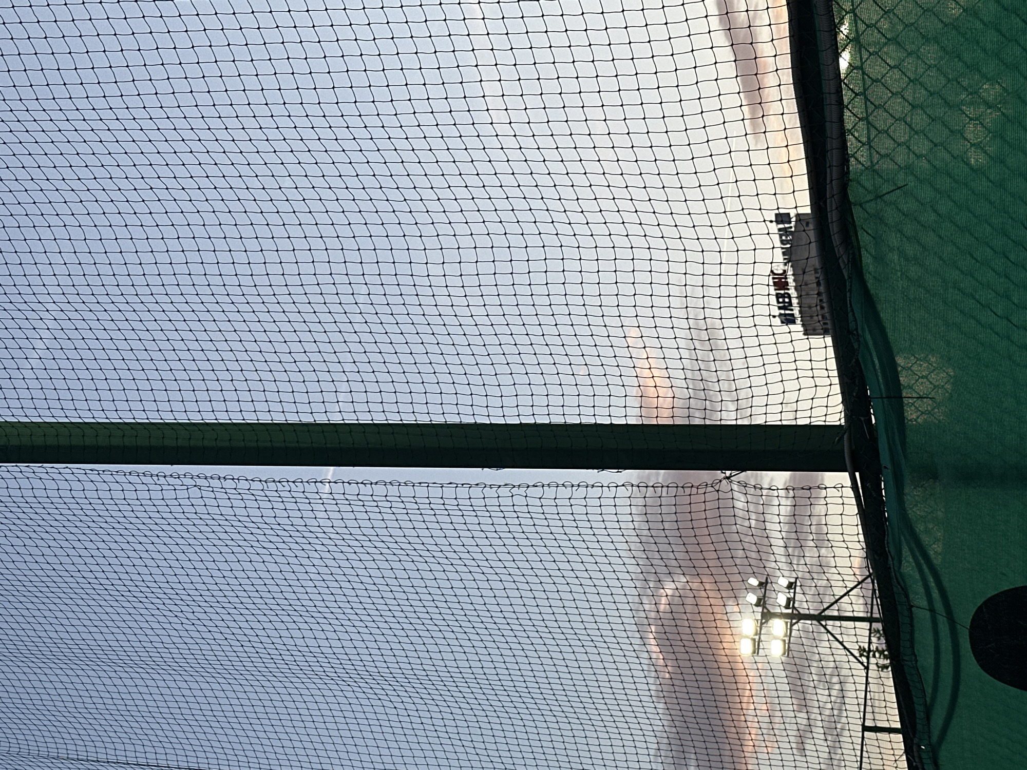 The image depicts a view through a black net, likely a sports fence, towards a dusky sky. A tall, dark green pole is visible on the left side, extending vertically through the frame. In the background, several bright stadium lights illuminate the sky, positioned above some vegetation. Further in the distance, a building with illuminated signage is visible. The sky displays soft hues of pink, orange, and blue, suggesting either sunrise or sunset. The foreground includes a lower green fence or barrier. The overall scene conveys an outdoor environment, possibly a sports field or stadium, in Sofia, Bulgaria, as indicated by the building's signage. No people are visible in the image.