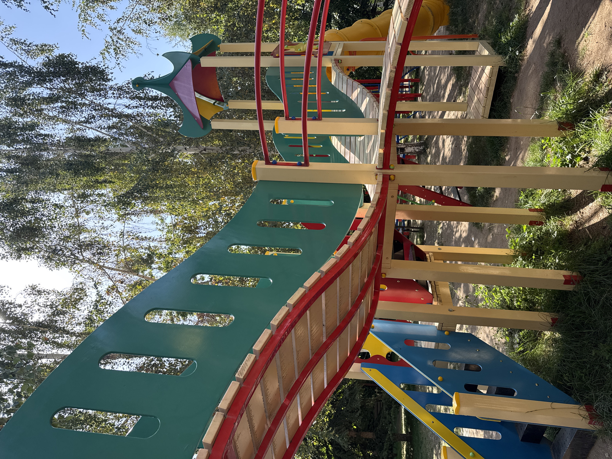 The image shows a colorful playground structure with green, red, yellow, and blue elements, set against a backdrop of trees and a bright blue sky. The playground features slides, ramps, and stairs, constructed from a combination of painted metal and wooden components. The ground beneath the structure is a mix of dirt and grass. The location is identified as Sary-Oy, Kyrgyzstan. There are no people visible in the image. The scene appears to be a sunny day.