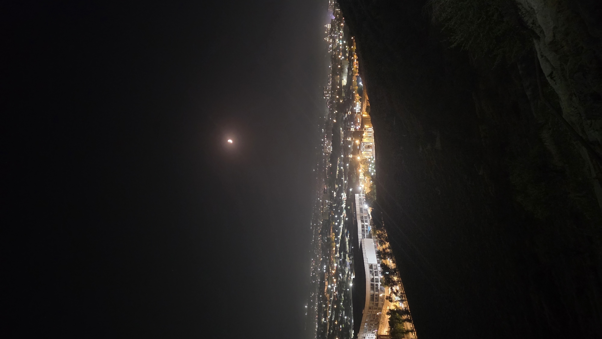 This is a nighttime, aerial shot of Osh City, Kyrgyzstan, taken from a high vantage point. The foreground is dominated by a dark, sloping hill covered in sparse vegetation, suggesting a natural, unlit area. In the midground, the city lights stretch out in a vast expanse, with numerous buildings illuminated by warm yellow and white lights. A prominent, modern-looking stadium with bright white lights is visible on the left side of the city. To the right of the stadium, a distinct, ornate building with arched entrances and golden illumination stands out. Trees are planted in rows along the edge of the hill in the midground, also lit by streetlights. The sky is dark, with a crescent moon visible in the upper right quadrant, casting a faint glow. The overall impression is a bustling city viewed from a peaceful, elevated natural setting at night.