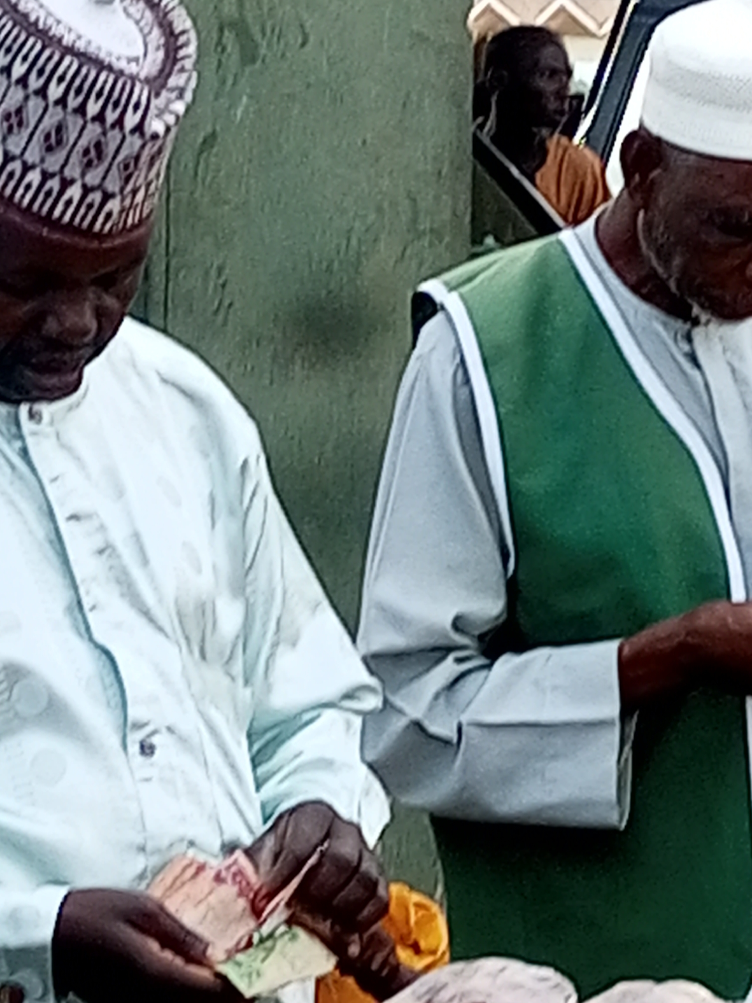 In Gombe, Nigeria, two men are captured in a close-up shot. The man on the left wears a patterned Kufi hat and a light blue shirt, his hands engaged in handling currency. To his right, another man is partially visible, wearing a white Kufi hat and a green vest over a light blue shirt. The background appears to be a textured green wall, with a faint glimpse of another person in an orange garment in the distance, possibly through an opening or doorway. The focus is on the immediate interaction between the two men, with the man on the left's hands and the currency he's holding being a prominent detail.