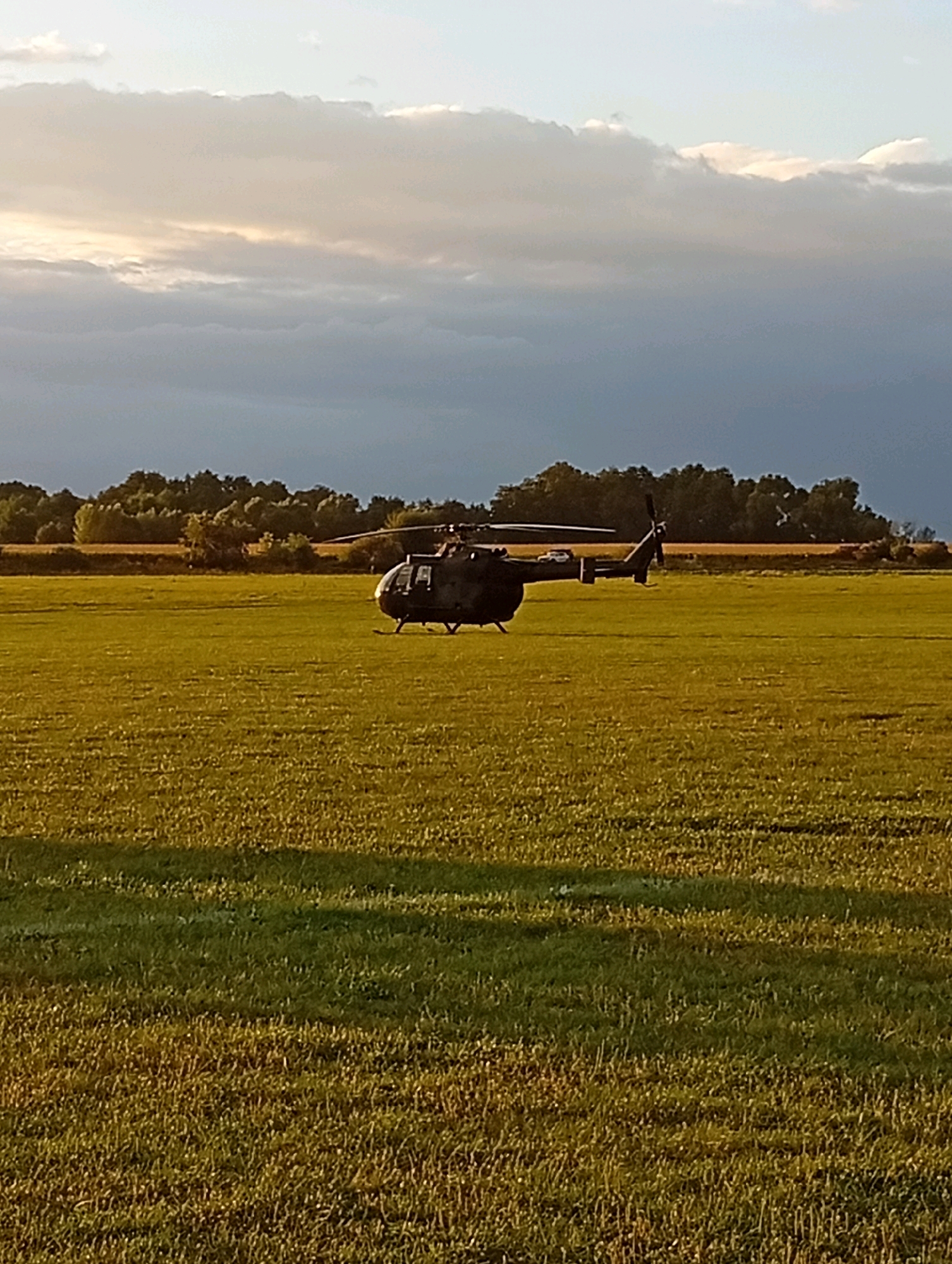 The image shows a military helicopter in a grassy field. The helicopter is dark green and appears to be on the ground, possibly having landed. In the background, there is a line of trees and a cloudy sky. The sun is low in the sky, casting long shadows across the field. The overall impression is one of a peaceful, rural setting. The image is undated and it is not possible to determine the exact location within Szewce, Poland.