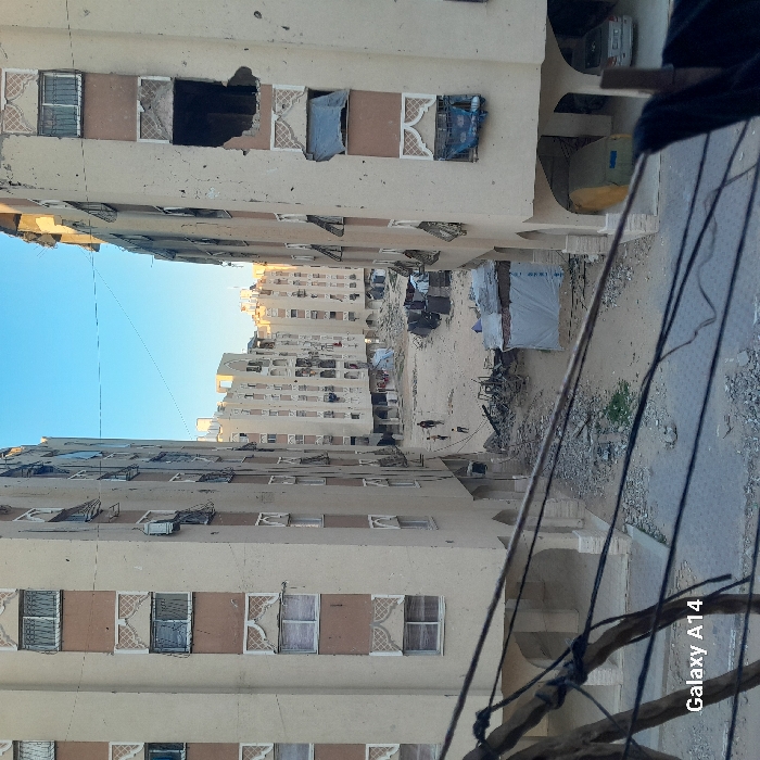 This image captures a wide view of a war-damaged urban environment in Khan Yunis, Palestinian Territory, looking down an open space between residential buildings. Two prominent multi-story apartment buildings flank the central area, both exhibiting extensive damage. Their facades, originally light beige with brown decorative bands and patterned white window surrounds, show numerous projectile impacts, shattered windows, and missing sections of wall; many window openings are covered with blue tarpaulins or fabric.
In the central ground area, a debris-strewn dirt path is visible, covered with rubble, twisted metal, and broken concrete. Several makeshift structures constructed from corrugated metal and tarpaulins are erected amidst the debris. Three small figures, appearing to be children, walk across this open space towards the background.
In the midground, additional apartment buildings, similar in design and also showing signs of damage, extend into the distance. The lower levels of the foreground buildings feature arched openings; one on the right reveals a yellow-covered vehicle and a silver sedan parked underneath. Overhead, multiple dark utility wires crisscross the foreground, partially obscuring the view. The sky above is clear blue.