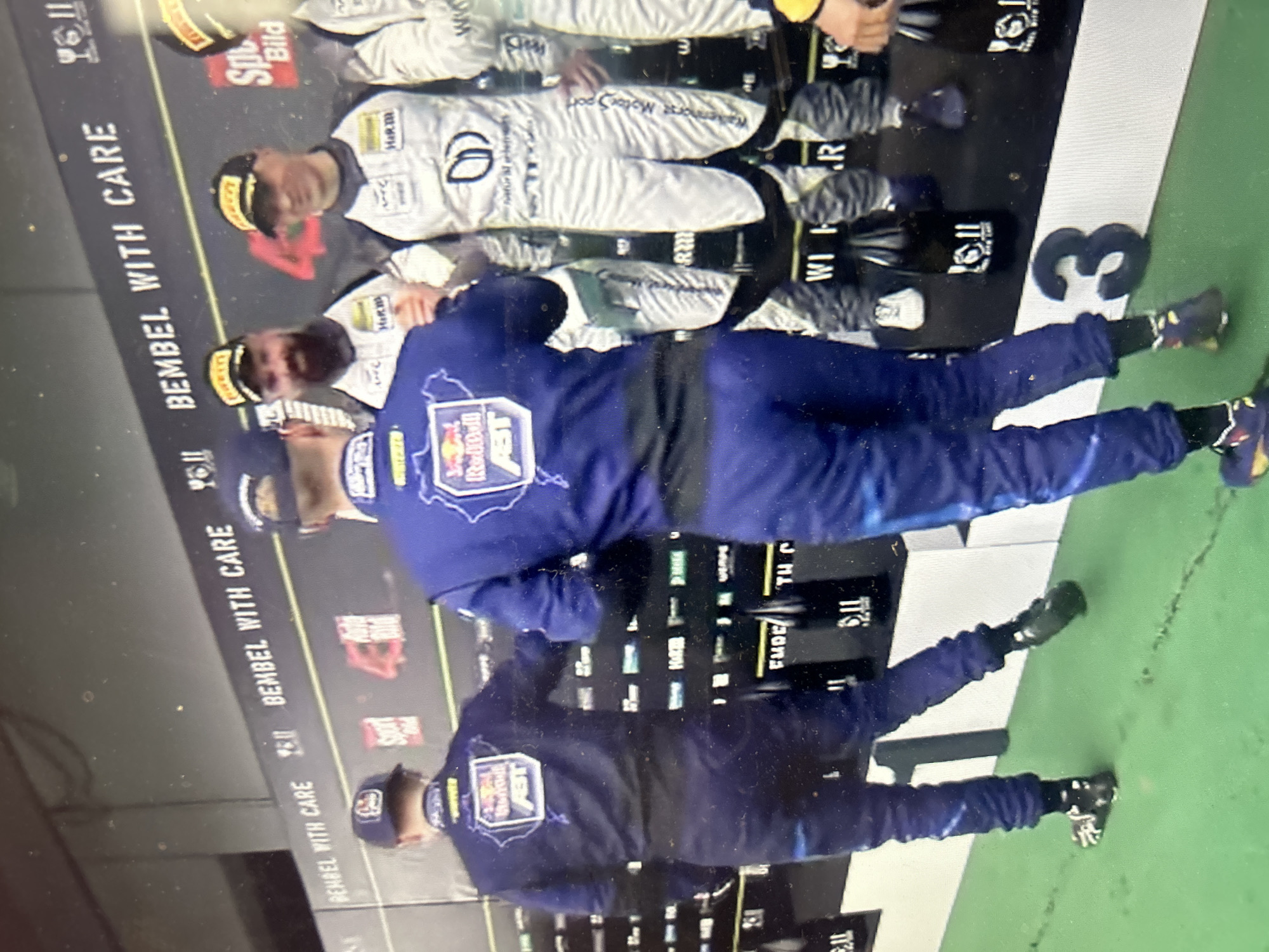 The image depicts a group of people on a podium. The podium displays "BEMBEL WITH CARE." One person wears a blue uniform with a "Red Bull ABT" logo and stands on the first position. Another stands on the second position in a white racing suit with "natural automotiv" and "Kalkenhorst Motorsport" written on it. Another stands in the third position in a white racing suit with "Wi Her" on it. The number "3" is visible on the podium. There is no information provided in the image or its OCR to indicate that the image was taken in Sofia, Bulgaria.