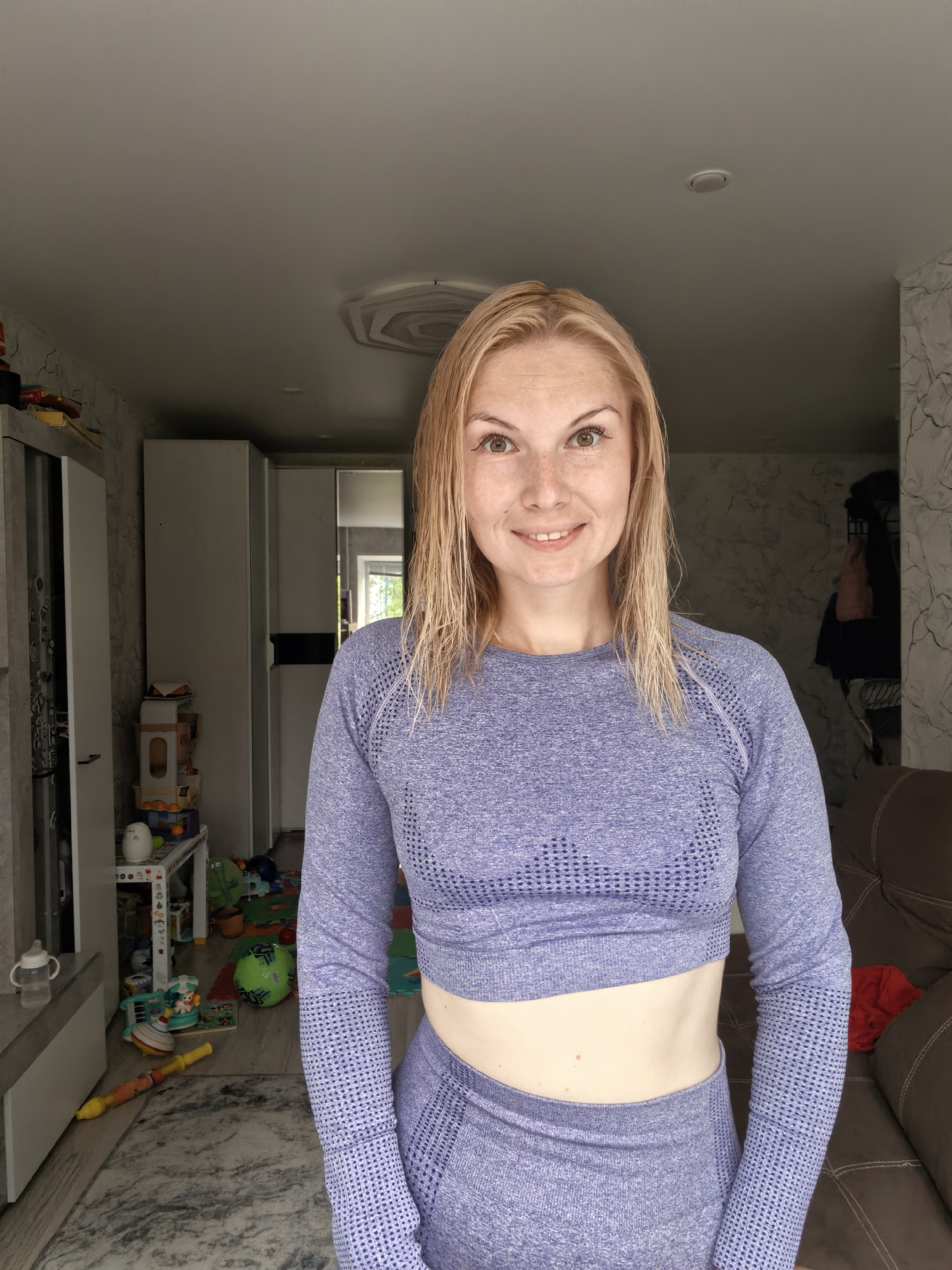 A young, fair-skinned woman with blonde hair and freckles, wearing a purple crop top and matching leggings, stands in a room with a cluttered background. The room appears to be a living area with children's toys scattered on the floor, a sofa in the background, and a wardrobe or cabinet against the wall. The woman is smiling directly at the camera. The overall impression is a casual indoor setting.