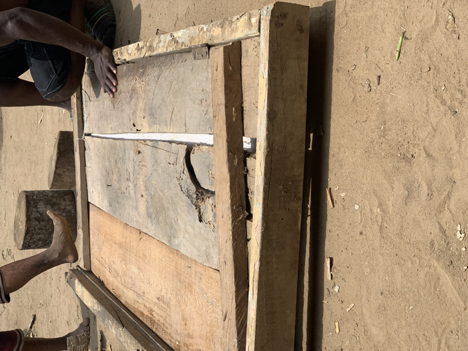 The image shows two people working on a wooden structure on a dirt ground in Egbeda, Nigeria. The structure is a large wooden frame made of several planks, which appear to be being joined together. One person is kneeling and using their hands to adjust the planks, while the other person's foot is resting on a large piece of wood that is propped against the structure. Small pieces of wood and sawdust are scattered around the structure on the ground. The scene is outdoors and the ground is sandy. The location based on the coordinates provided is in the suburb of Egbeda in Lagos, Nigeria.