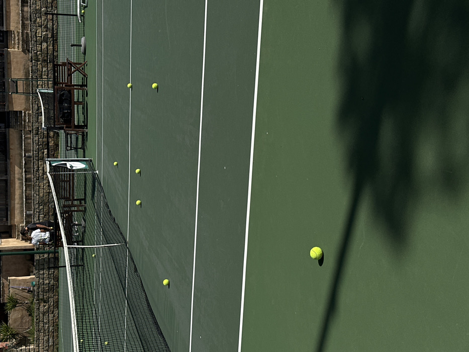 A forensic analysis reveals an outdoor tennis court situated in Ericeira, Portugal. The court features a dark green hard surface demarcated by white lines and a black mesh net with a white top tape and a green and white branded side strap on the right. Eleven yellow tennis balls are visible across the court, scattered on both sides of the net, with one ball prominent in the lower foreground. In the background, two individuals stand behind the net; one wears a white shirt and dark trousers, the other a dark shirt and dark trousers. Behind them, a stone retaining wall supports a level ground featuring two dark wooden benches and portions of a light-brown, possibly terracotta-colored, building. Strong sunlight illuminates the scene, creating sharp shadows from the net, balls, and other unseen elements to the lower right.