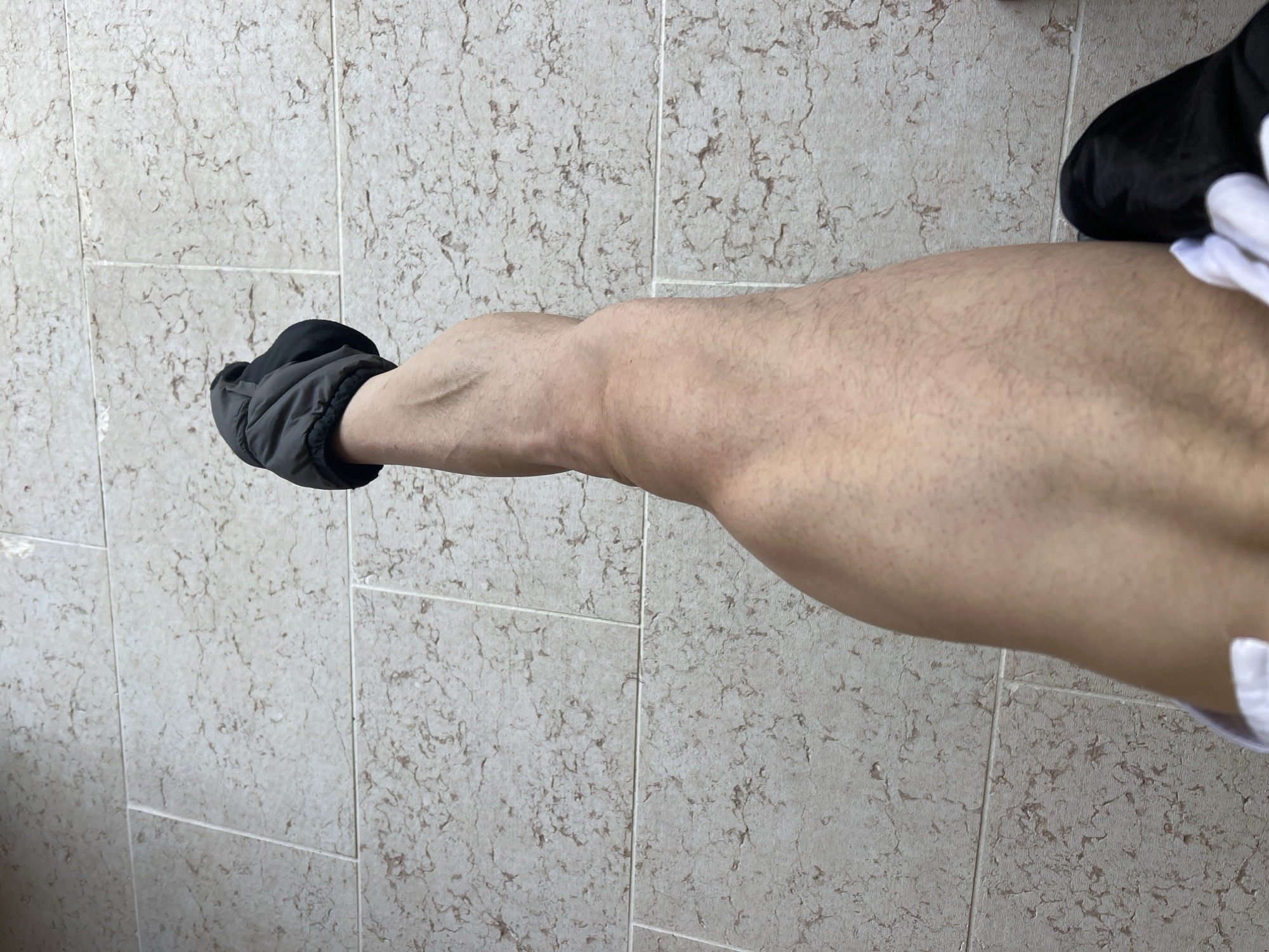 The image shows a person's right leg and foot, which is clad in a black, quilted slipper. The leg is mostly bare, revealing a tanned complexion and visible muscle definition, particularly in the calf. The foot is pointed upwards, suggesting the person is standing or sitting with their leg raised. The background consists of a tiled floor with a light beige color and a natural stone pattern. The tiles are large and rectangular, laid out in a grid pattern with visible grout lines. The lighting appears to be natural, casting soft shadows. The scene is devoid of any specific landmarks or indicators that would place it in Verona, Italy. The image is a close-up shot, focusing solely on the leg, foot, and flooring.