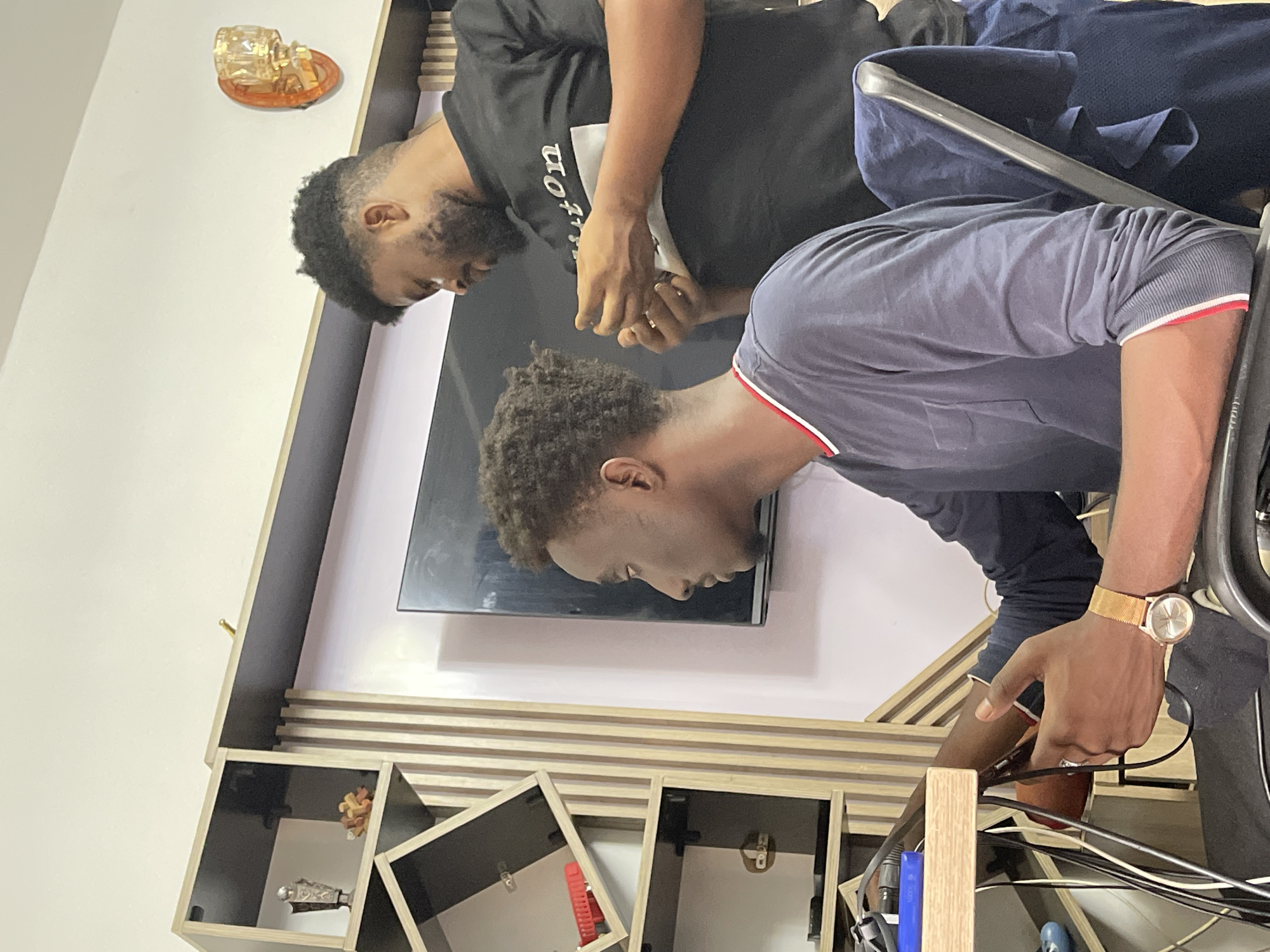 Two men are present in an indoor setting, likely a residential space given the visible furniture. One man is wearing a black t-shirt and appears to be focused on something in his hands. The second man is wearing a dark blue long-sleeve shirt with a red and white trim on the sleeves and is working with cables near a wooden cabinet unit. The cabinet unit has shelves with dark interiors and some items are stored within. A television screen is mounted on the wall above the cabinet. The scene may be set in Abuja, Nigeria, although there is no explicit evidence in the image to confirm this definitively.