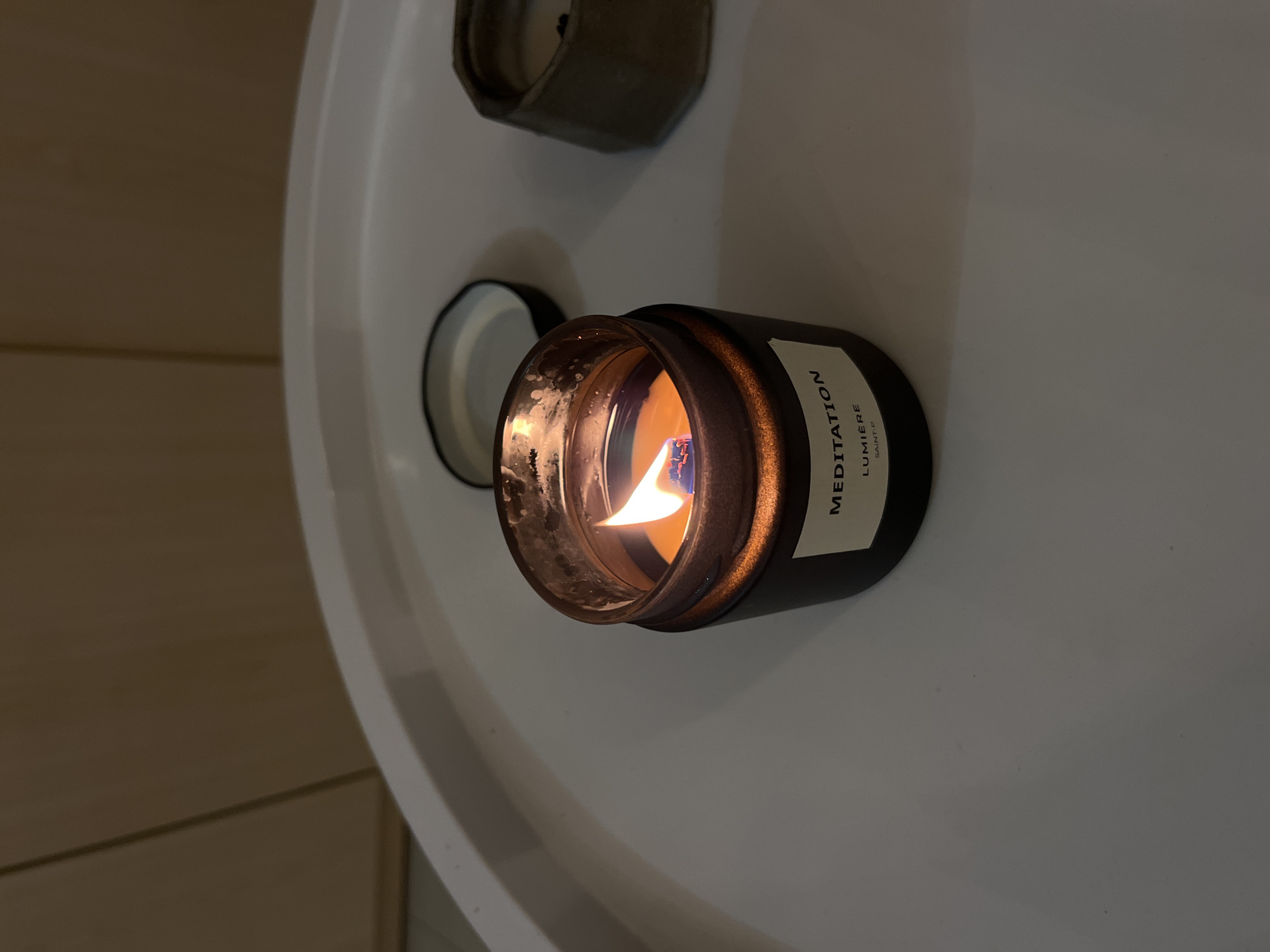 A lit candle with a bronze-colored container and a white label that reads "MEDITATION LUMIERE SAINT-P" is positioned on a white surface. Next to the candle is its lid and another dark green candle in a similar container. The setting appears to be indoors on a white table, with a wooden wall visible on the side. The location is not discernible as being Sochi, Russia, based solely on the visual information provided in the image.