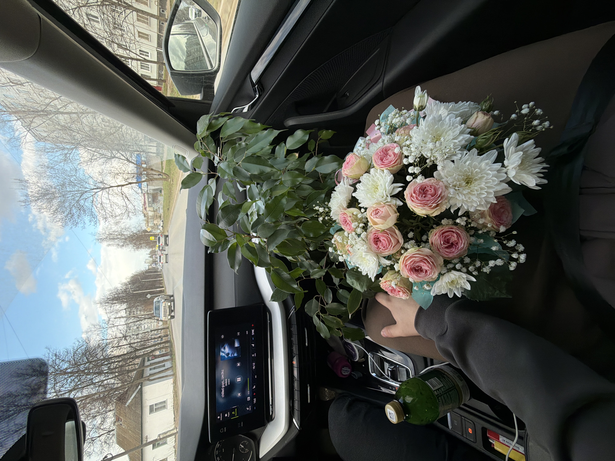 A first-person perspective image from inside a car captures a large, vibrant bouquet of flowers held by a person in the passenger seat, with a street scene visible through the windows. The image is oriented vertically.
In the foreground, a person's hand, wearing a dark long-sleeved top, supports a lush bouquet. The bouquet prominently features pink roses, white chrysanthemums, and delicate white filler flowers (like baby's breath), intertwined with abundant green foliage and wrapped in light blue and brown paper. To the right of the bouquet, a green bottle with a yellow cap rests in the car's center console. On the dashboard, a digital infotainment screen displays music information, showing "chilled guys, Lin3kz" as the current track by "Undore." Car interior elements such as the door panel, handle, and rearview mirror are also visible.
Through the car's windows, a daytime street scene unfolds under a bright, partly cloudy sky. Bare trees line the road, suggesting late winter or early spring. Various buildings, including multi-story residential blocks and smaller houses, are visible. A white SUV is seen driving further down the road. The architecture and streetscape are consistent with the provided context of Chaikovsky, Russia. The scene conveys a moment of travel or pause, with the flowers being a central focus.