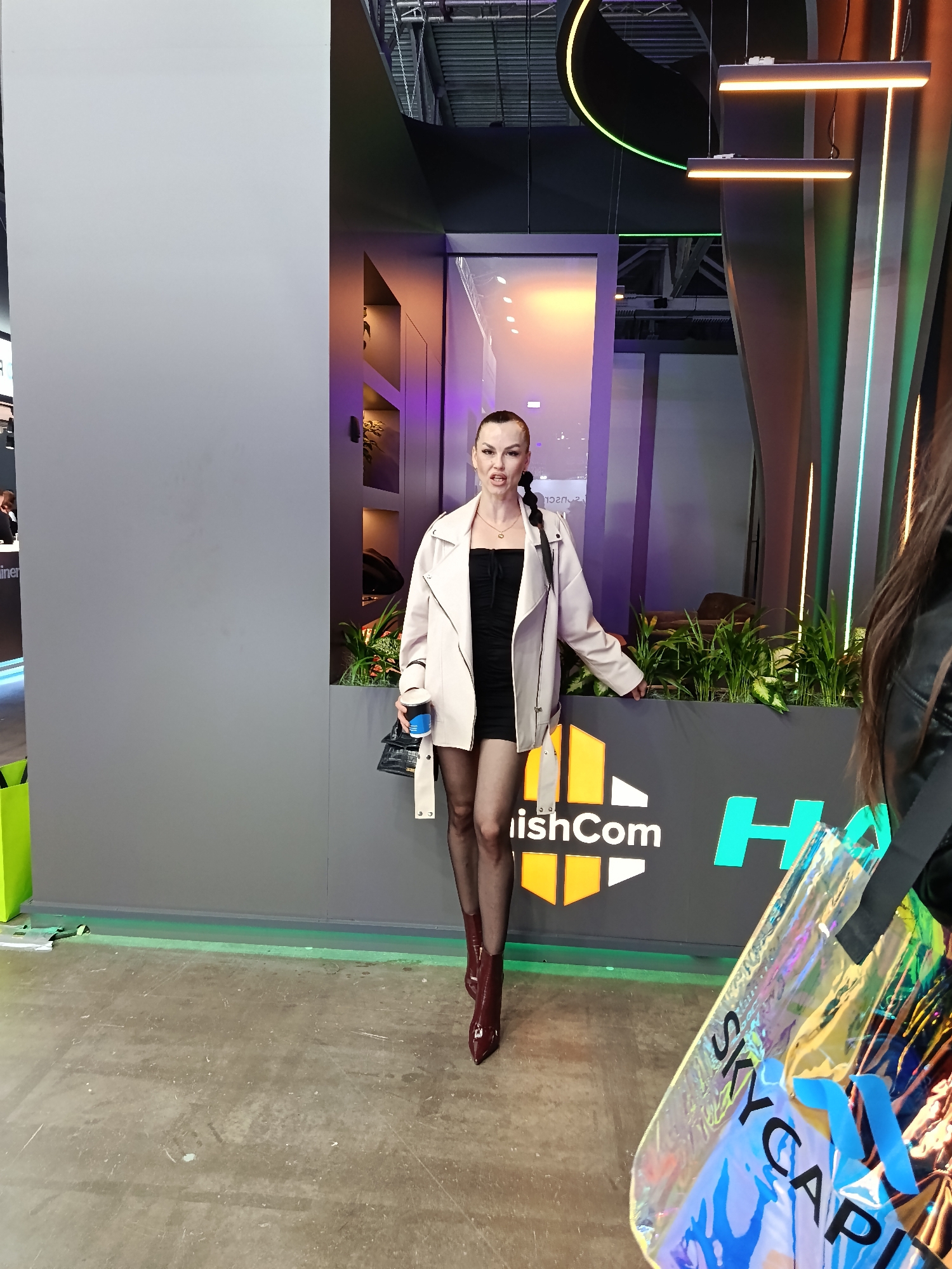 The image shows a woman standing in front of a booth, likely at a conference or trade show, in Moscow, Russia. The woman is wearing a black dress, fishnet stockings, and burgundy boots, with a light-colored jacket. She has her hair in a braid and is holding a drink in her left hand. The booth behind her has a sign that says "ishCom," and there are plants on top of the sign. The background includes modern architectural elements with neon lighting. A second person is partially visible on the right, holding a "SKY CAPITAL" bag. The floor is concrete.
