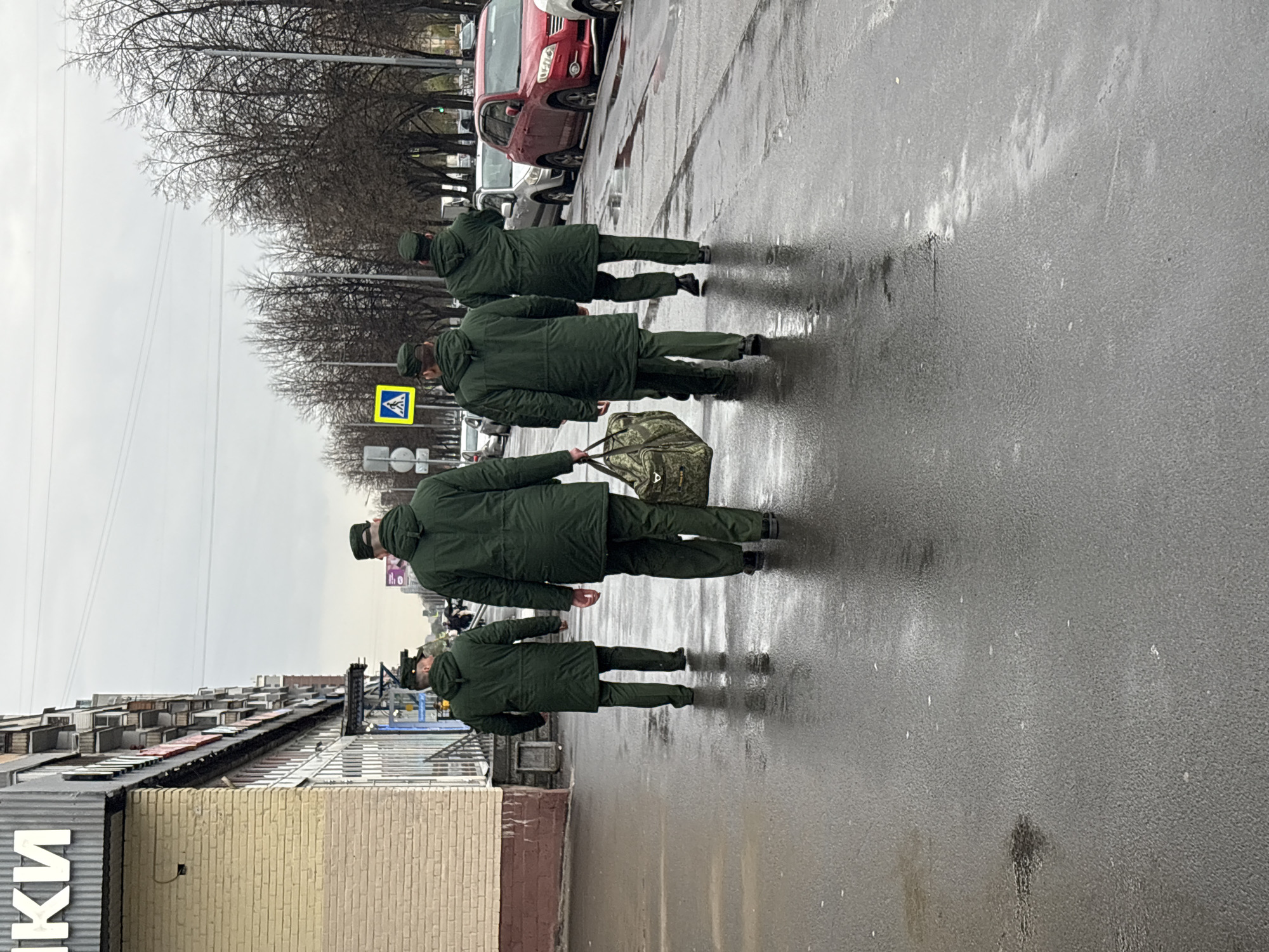 Four individuals in matching green military uniforms walk in single file on a wet street. The rear individual carries a duffel bag. The setting appears to be an urban environment, as suggested by the presence of parked cars, buildings, and street signs. Bare trees line the street. The overall mood is somber due to the overcast sky and the serious nature of the individuals walking. The location is specified as Novoe Devyatkino, Russia.