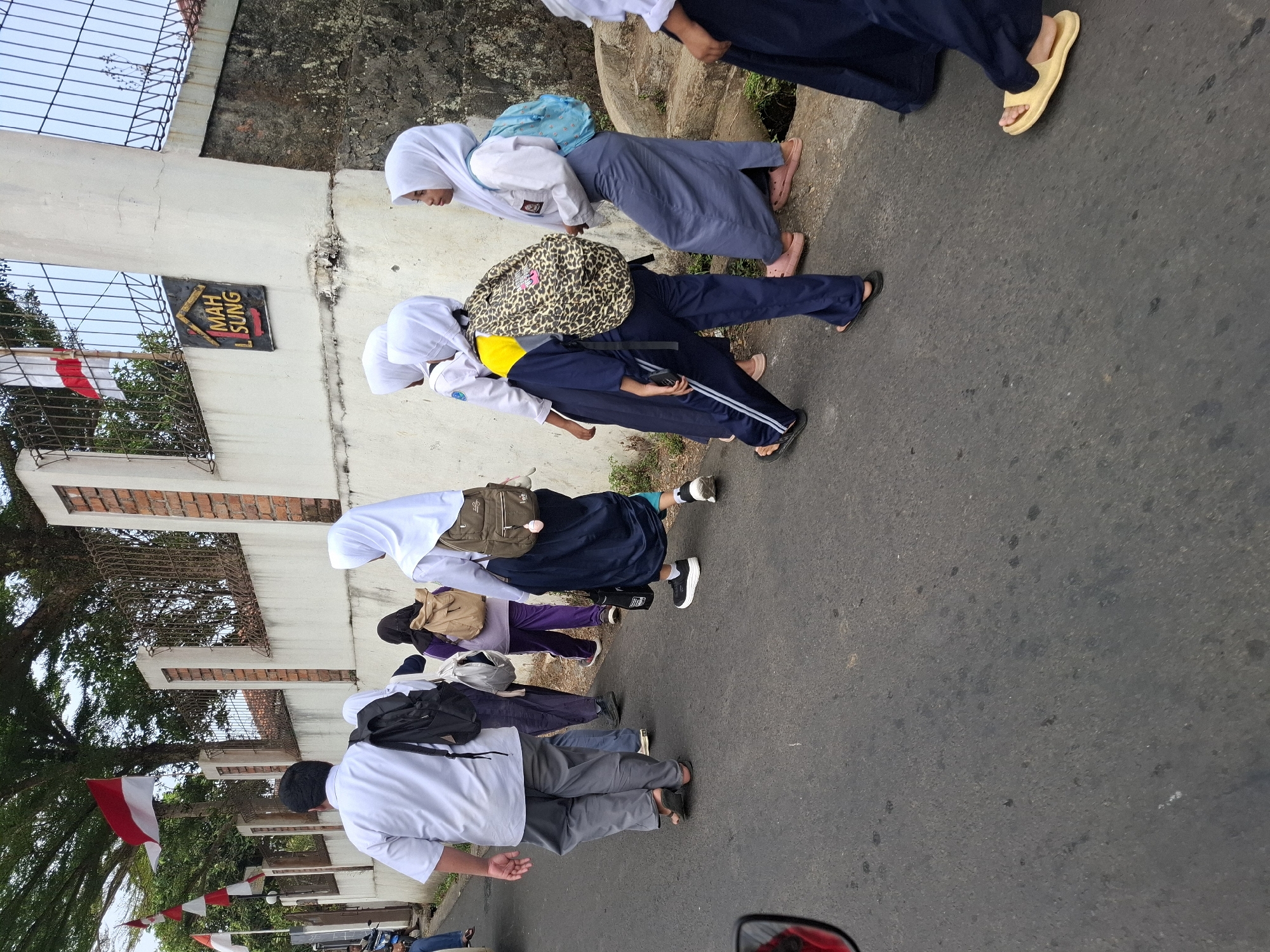 A group of students wearing school uniforms are walking on a paved road in Balekambang, Indonesia. The students, some wearing hijabs, are carrying backpacks. In the background, there is a white wall with a sign that reads "MAH LISUNG" and Indonesian flags are displayed. The weather appears to be overcast.