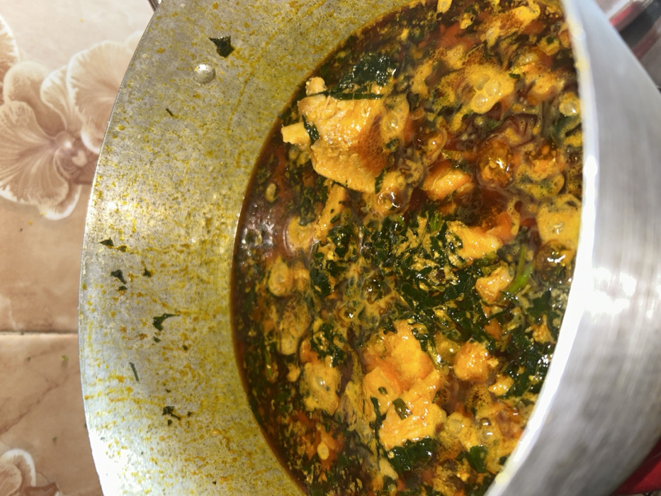 The scene depicts a large, metallic cooking pot containing a prepared, simmering dish, located in Egbe, Nigeria. The pot holds a robust stew or soup, characterized by a vibrant reddish-orange oil base with visible floating oil droplets. Within the liquid, distinct elements of dark green leafy vegetables and irregularly shaped, light orange-yellow protein pieces are present. The inner rim and upper walls of the pot show extensive residue and splatters from the cooking process. The immediate background consists of a tiled wall featuring light brown or beige square tiles with an embossed, subtle floral or shell-like pattern. No individuals are present, and no actions or interactions are depicted. The environment is consistent with an indoor kitchen setting.