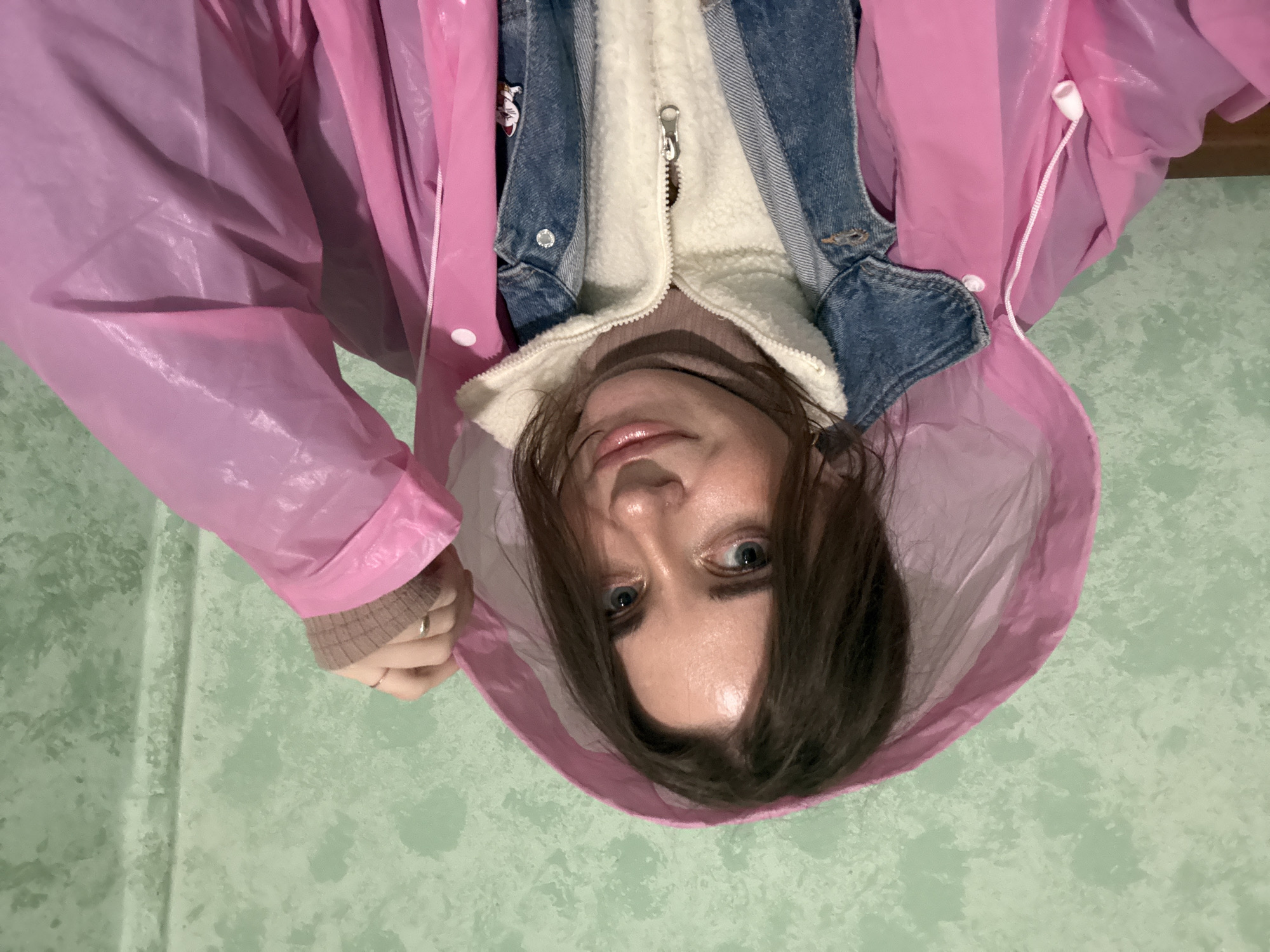 A woman is looking down at the camera while wearing a pink, plastic raincoat, a denim jacket, and a beige fleece jacket. She is indoors with a textured, light green wall in the background. The location is not identifiable as Moscow, Russia from the visual information provided.