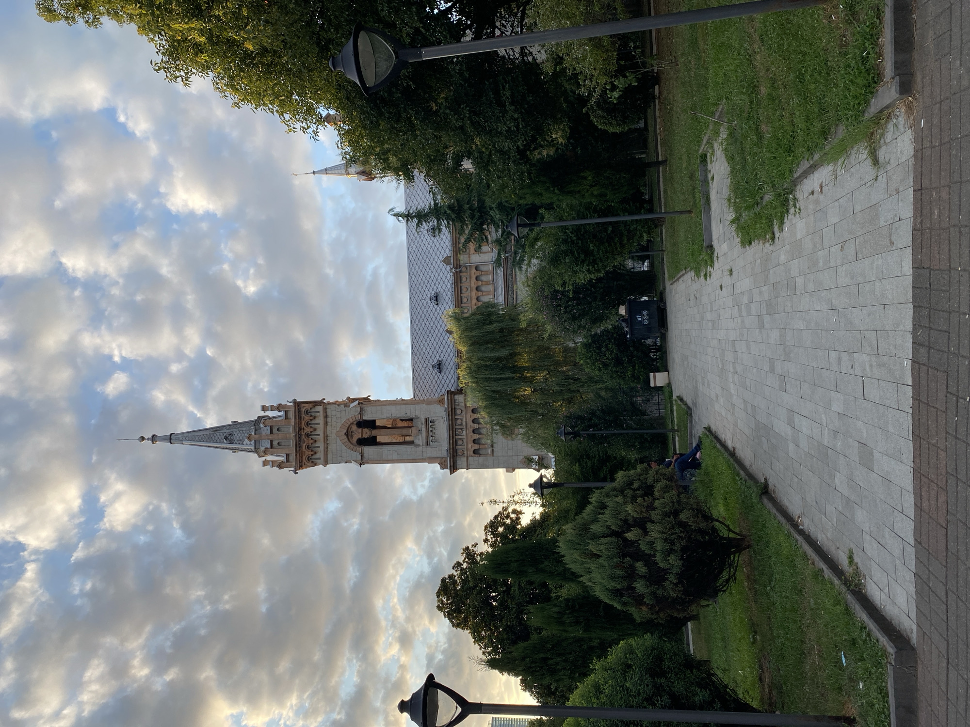 The image depicts a view of the Holy Spirit Catholic Church in Batumi, Georgia, under a cloudy sky. A paved pathway leads through a grassy park area with trees and bushes towards the church, which has a prominent tower with a spire. Several lampposts are visible along the pathway. In the mid-ground, a person is seated on a bench in the park. A trash receptacle is also present. The overall scene suggests a public park or square adjacent to the church.