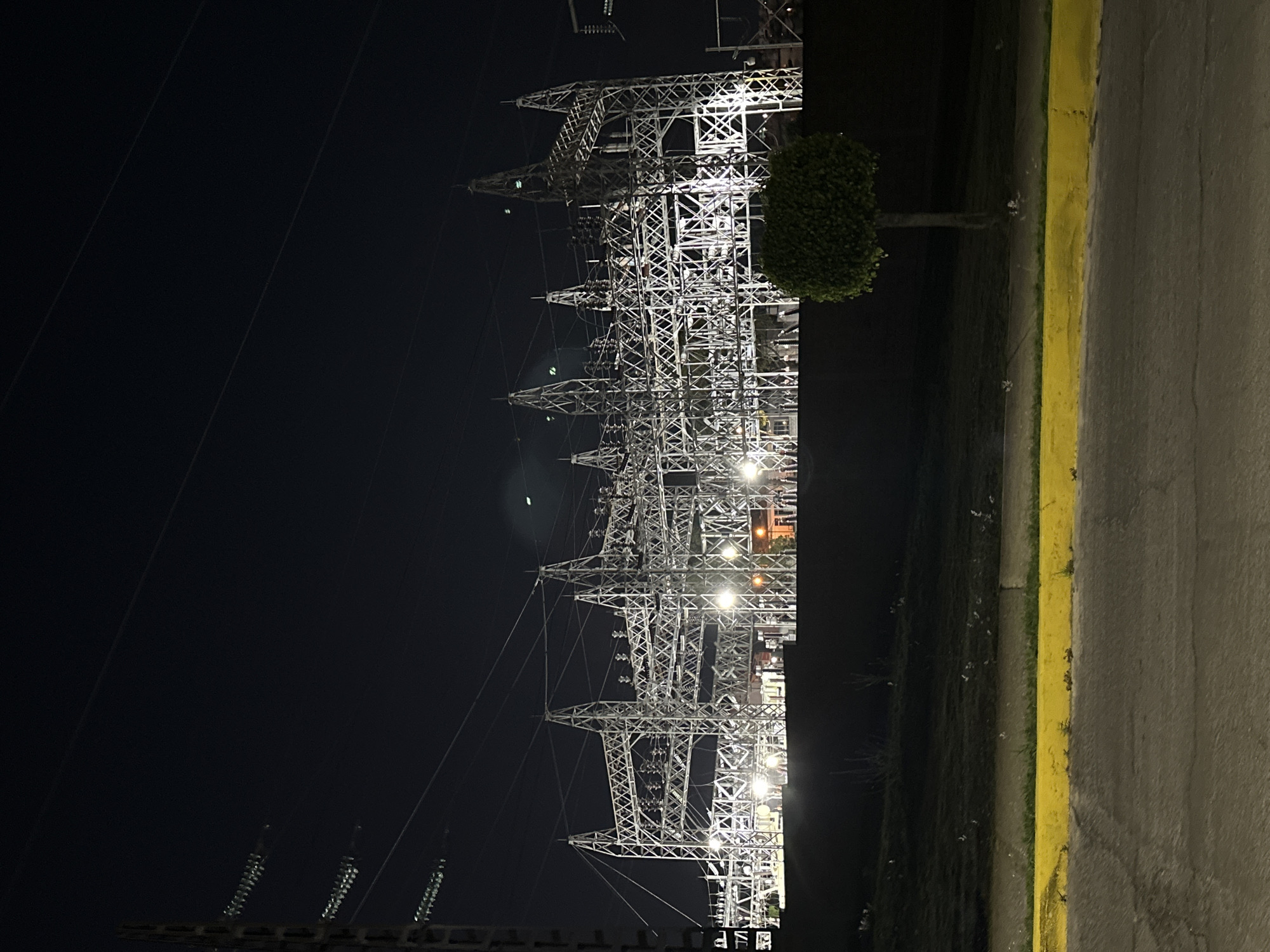 At night in Puebla, Mexico, a power substation stands illuminated against a dark sky. The substation's metal framework is intricate and tall. Power lines stretch out into the darkness. A small, round tree sits near the edge of the frame, partially obscuring the substation. A concrete sidewalk bordered by a yellow line runs along the edge of the image.