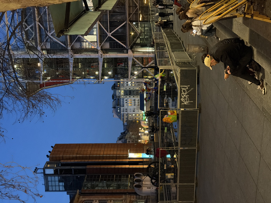 The image depicts the Place Igor Stravinsky in Paris, France, at twilight or early evening. The Centre Pompidou, identifiable by its exposed white structural framework, exterior escalators, and utility pipes, dominates the right side of the background. To the left, a tall, rectangular building with dark glass and orange-brown paneling stands against a deep blue sky.
In the mid-ground, a section of the plaza, including the Stravinsky Fountain (also known as the Fontaine des Automates), is cordoned off by grey metal construction barriers. Several colorful, abstract sculptures from the fountain, including a prominent red heart and a multi-colored figure, are visible within the fenced area, which contains water. Two individuals in high-visibility yellow-orange vests are situated inside the barriers, suggesting ongoing work or maintenance. Graffiti spelling "KSRGU" is present on one barrier. Further left, within the fenced area, a white industrial-looking structure with three large circular vents is visible.
In the foreground, a paved area leads to a set of steps where multiple individuals are seated. On the lowest step, a person wearing a beige baseball cap and a dark jacket is seated, holding a cigarette or small object. Adjacent to them, another individual in a white top is partially visible. Further right, a group of approximately 4-5 people are seated on the steps, some utilizing yellow wicker-style cafe chairs, with at least one person looking at a mobile phone. A dark cylindrical receptacle stands near the seated group. Beyond the barriers, near the base of the Centre Pompidou, a small group of 4-5 individuals are standing on the pedestrian area. Bare tree branches are visible in the upper frame against the twilight sky. Distant Parisian buildings with illuminated windows are visible in the far background.