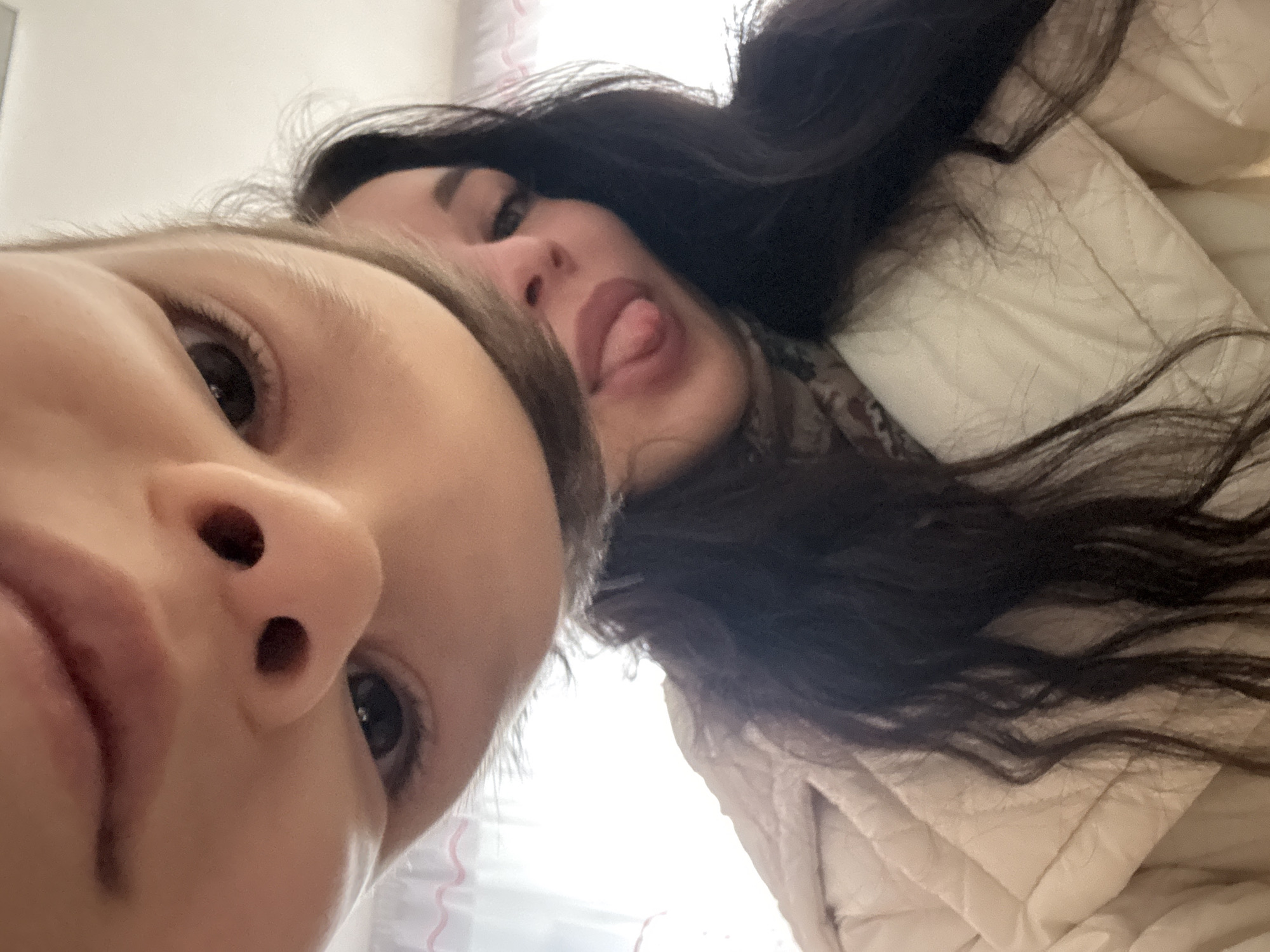 Two individuals are captured in a close-up, selfie-style image. The foremost figure is a child with fair skin and light-colored hair. Their face dominates the lower left quadrant of the frame, with features like a prominent nose and visible eye. Positioned behind and to the right is an adult woman. She has long, dark hair and is sticking her tongue out, adding a playful element to the image. The background appears to be an indoor setting, with a beige, possibly quilted material partially visible behind the woman. No specific details are available regarding the precise location within Chaikovsky, Russia.