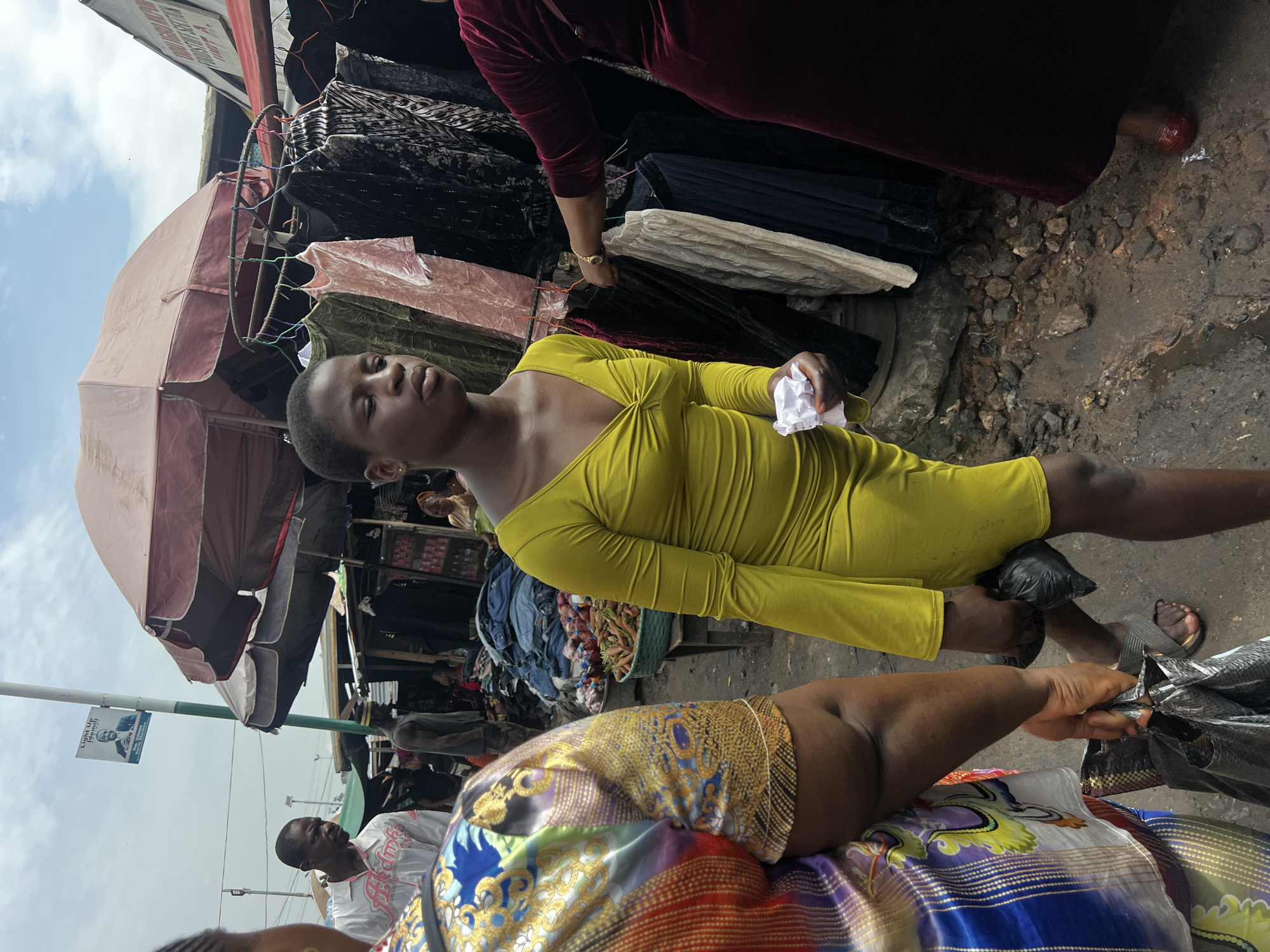 In Nkpor, Nigeria, a young woman in a bright yellow dress walks through a market. She holds a crumpled piece of white paper in her left hand and a black plastic bag in her right. Behind her, a woman in a vibrantly patterned traditional outfit carries another black plastic bag. In the background, stalls display various goods, including clothing on hangers and baskets of produce. The ground is uneven and appears to be dirt with some puddles, indicating a potentially dusty or muddy environment. A man in a white shirt with text is visible further in the background, and a political poster is attached to a lamppost overhead. The scene suggests a bustling marketplace, characteristic of urban Nigerian life.