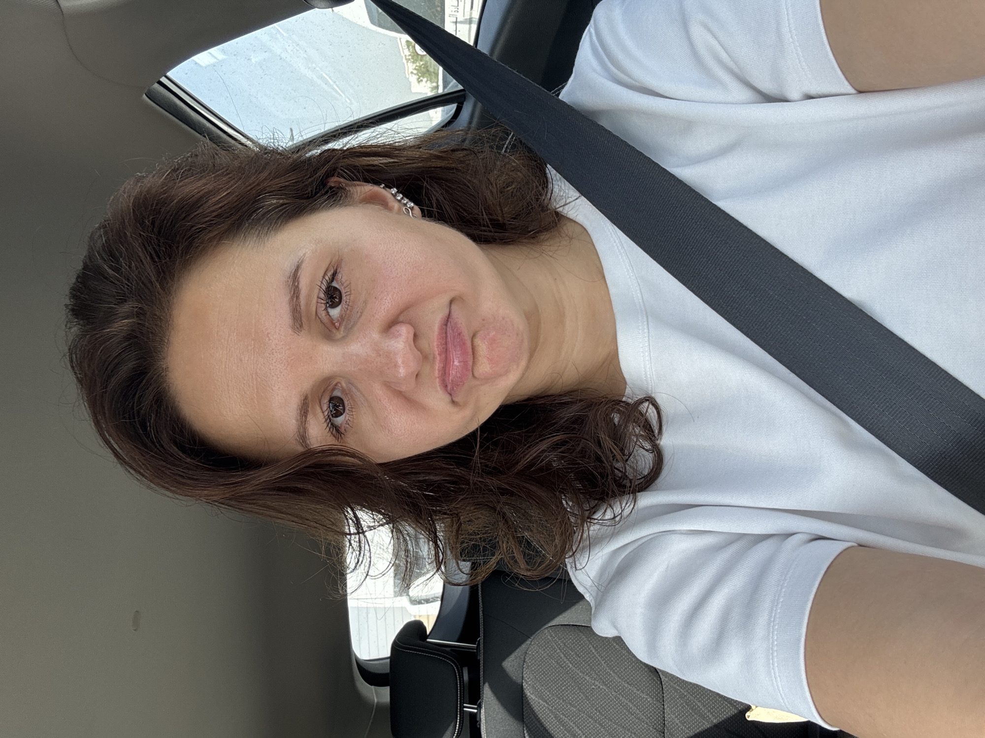 A woman with brown hair and brown eyes is seated in a car, wearing a white shirt and a seatbelt. The interior of the vehicle is visible, along with a partial view of the outside through the car window. The location cannot be determined to be in Dubai, United Arab Emirates, based on the visual information provided.