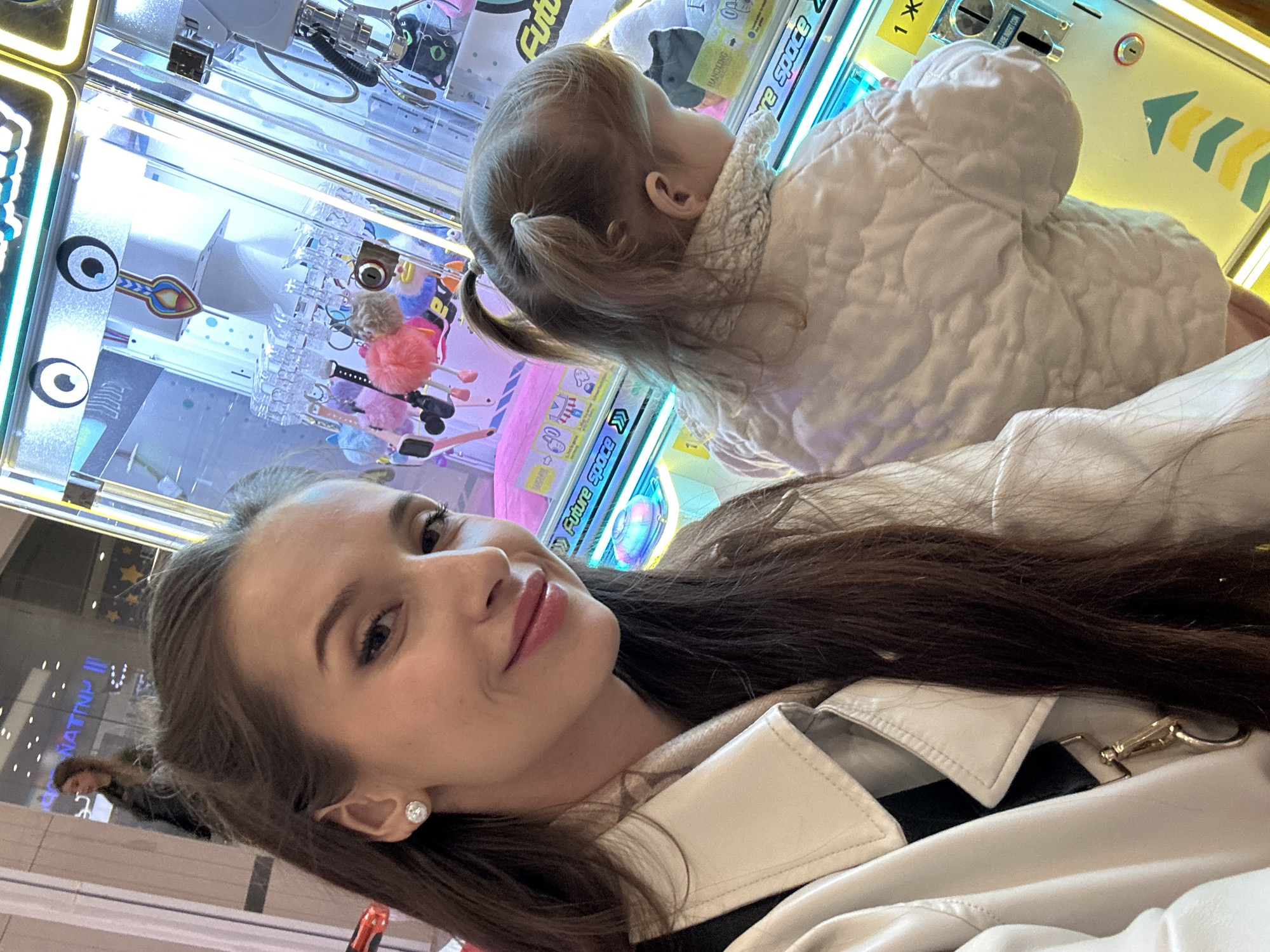 A selfie of a woman with long brown hair and an earring is in the foreground, while a small child in a white quilted jacket faces a claw machine filled with toys. The woman is wearing a light-colored jacket with black trim. The claw machine has "Future Space" written on it. The photograph appears to be taken indoors, possibly in a shopping mall. In the background, one other person is partially visible. From the text on the claw machine, the photo was taken in Yekaterinburg, Russia.