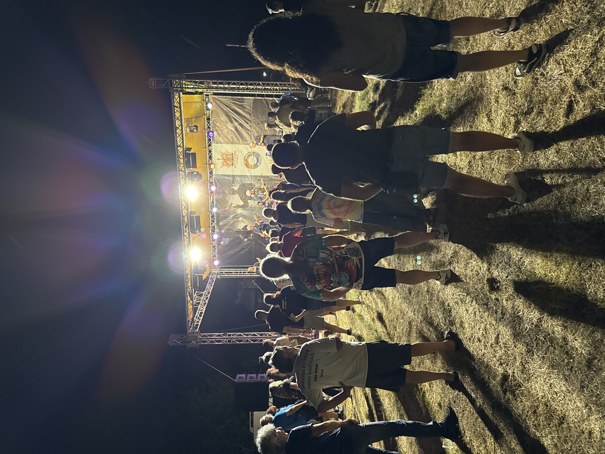 The image shows an outdoor concert at night, possibly in the city of Tutrakan, Bulgaria, given the context. A crowd of people are standing on dry, grassy ground, facing a brightly lit stage. The stage has a large banner behind the performers, featuring a logo and graphics. Several band members are visible on stage, playing instruments and singing. The lighting on stage is a mix of bright spotlights and colored lights, casting beams into the night sky. Lens flares are prominent in the upper portion of the image due to the stage lights. The crowd appears engaged, with some individuals holding up phones. The overall atmosphere suggests a lively musical event.