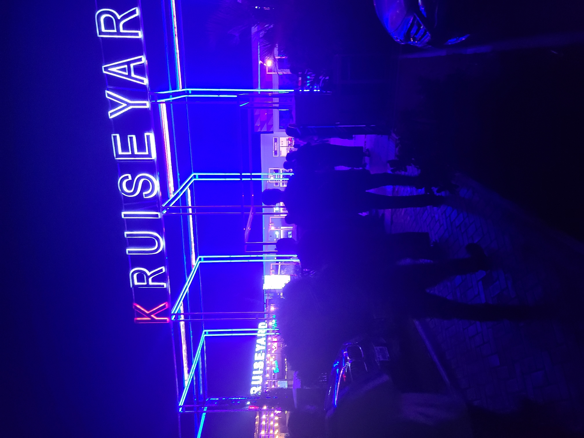 The image shows a night scene at "KRUISEYAR" (presumably a business or location name) in Gura Top, Nigeria. The name is displayed vertically in large, illuminated neon letters and bordered by blue neon outlining. In the mid-ground, silhouettes of several people standing near the establishment are visible, partially obscuring the background. The background features other buildings with illuminated windows and signage. A car is parked in the foreground. The scene is dominated by blue light.