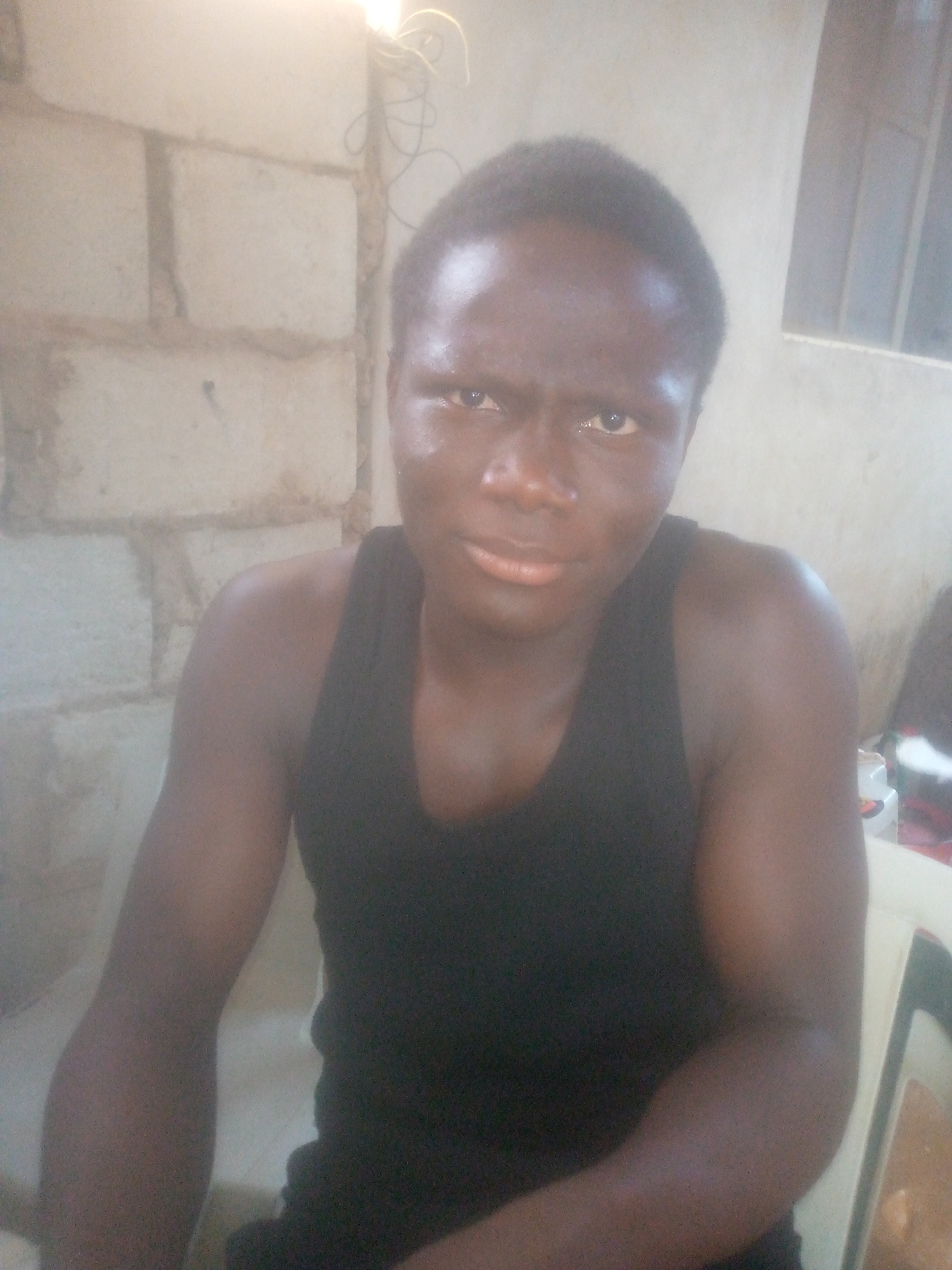 A medium shot of a dark-skinned man wearing a black tank top. He is seated. Behind him is a wall constructed of cement blocks. To the right is a window and what appears to be a plastic chair. Above the man's head are wires hanging. The location is Unknown, Unknown.