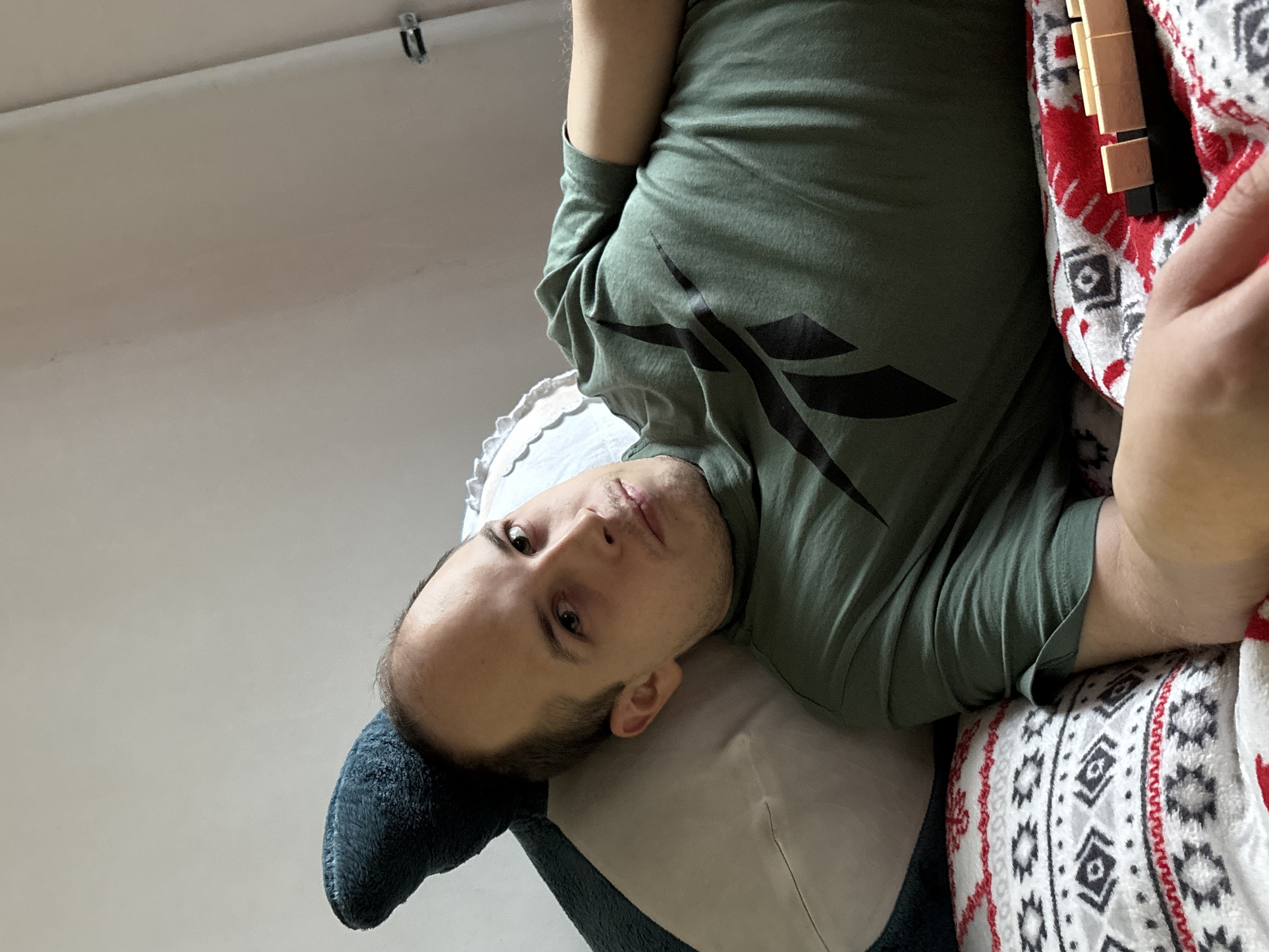 A man is lying on a pillow with a blanket over him. He is wearing a green t-shirt with black markings on it. The environment appears to be indoors. The location is not discernible from the image.