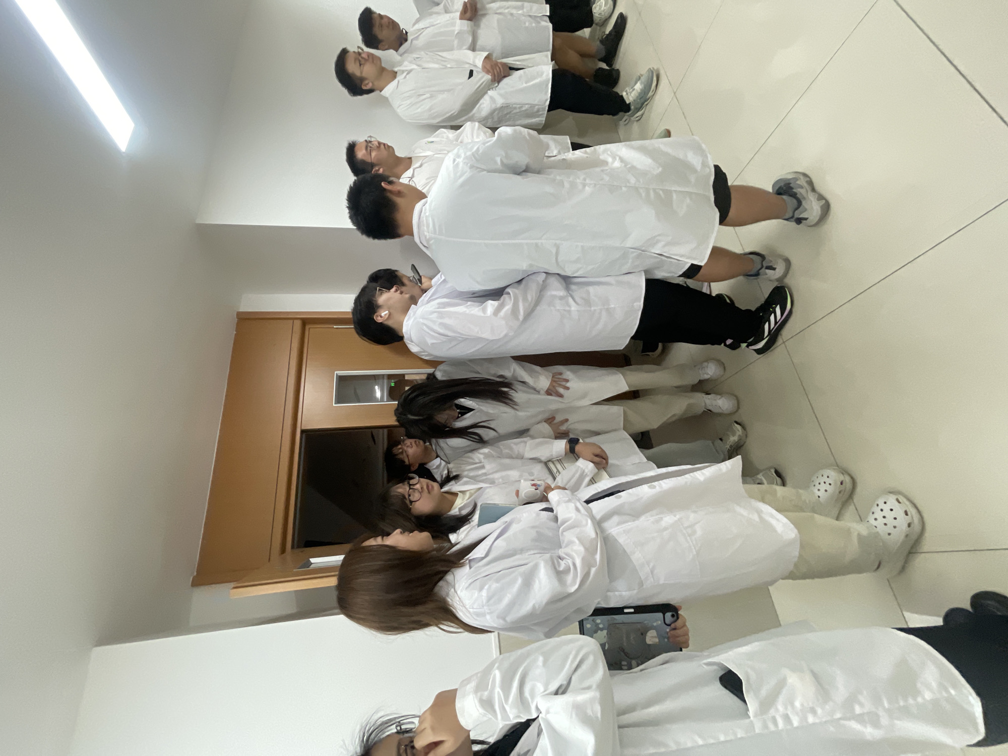 A group of approximately ten people, most wearing white lab coats, stand in a hallway. The hallway appears to be indoors, possibly in a laboratory or educational facility, based on the attire. The walls are white, and a light fixture is visible on the ceiling. A wooden door is situated to the left of the frame, with another doorway visible in the background. Several individuals are looking in the same direction, suggesting they are observing or listening to something. One person in the foreground on the right is facing away from the camera. The lighting suggests it is daytime. The image is taken from a low angle, focusing on the lower bodies of some individuals and the midsections of others. The floor is tiled with light-colored square tiles.