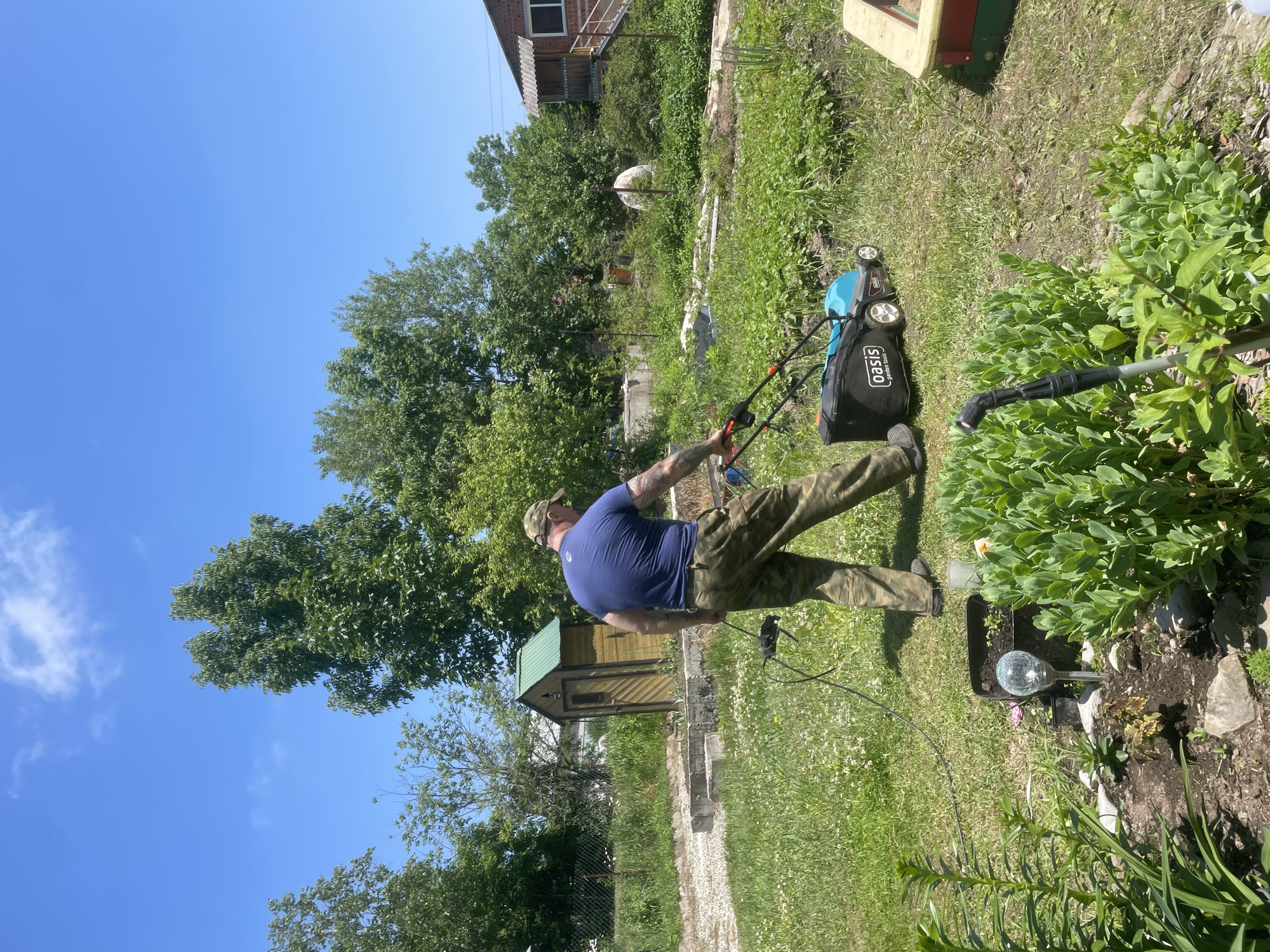 A man wearing a blue t-shirt and camouflage pants is mowing a grassy lawn with a light blue and black lawnmower. He is wearing a camouflage baseball cap. The lawnmower has the brand name "Oasis" visible on it. He is standing in a yard with trees and bushes in the background. There is a small wooden shed with a green roof behind him. A brick house with a balcony is visible to the right of the image. The sky is clear and blue. The scene appears to be a residential area.