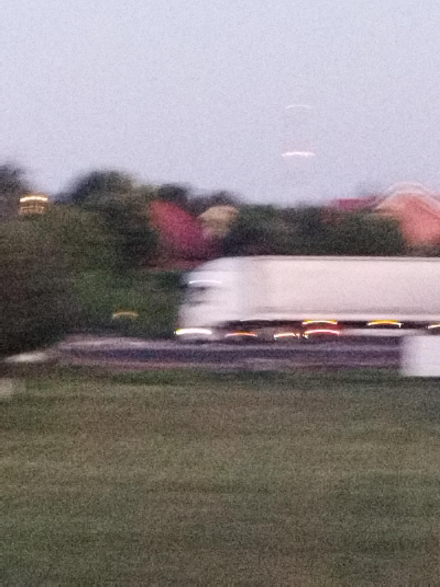The image, likely captured in Bucharest, Romania, depicts a blurry, low-light scene focused on a semi-trailer truck in motion. The truck, white with a long trailer, is the primary subject, its movement indicated by streaks of light from its taillights and possibly headlights. The trailer appears to be traveling on a road or highway, suggested by the blurred linear structures beneath it, possibly a median or lane markers.
The background is a soft, indistinct landscape. To the left, a dense cluster of trees or bushes is visible, with a faint, warm light emanating from within, hinting at buildings or structures obscured by the foliage. Further into the background, abstract shapes of red and pink suggest rooftops or structures, possibly houses or commercial buildings. The sky is a muted, overcast grey.
In the distance, at the top of the frame, two faint, elongated white streaks are present in the sky, possibly reflections or lens flares, though their exact nature is unidentifiable due to the image quality. The foreground consists of a grassy area, also slightly blurred, indicating the camera was positioned at a moderate distance from the truck, possibly from the side of the road or a nearby elevated position. No people are discernible in the image. The overall impression is one of movement and an urban or suburban periphery at dusk or dawn.