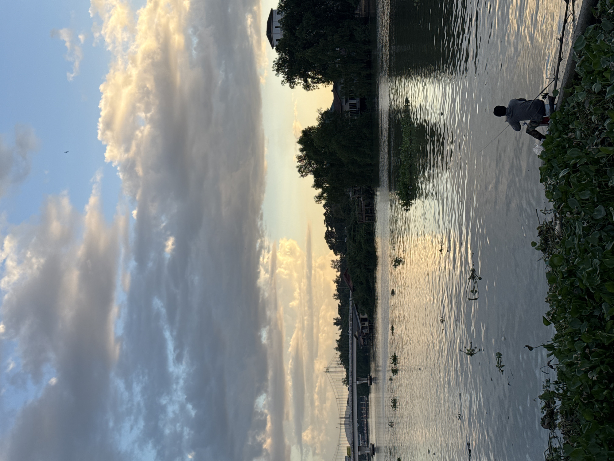 The image depicts a serene riverside scene at dusk in Surat Thani City Municipality, Thailand. In the foreground, lush green vegetation lines the riverbank, with a person seated on a log, fishing with a rod. The river's surface reflects the soft, warm hues of the setting sun and the dramatic, cloud-filled sky. Across the river, a dense line of trees obscures the view of buildings, with a prominent white structure visible atop a hill. In the distance, a long bridge spans the river, with cars visible on its roadway. The overall atmosphere is one of tranquility and calm, characteristic of a peaceful evening by the water.