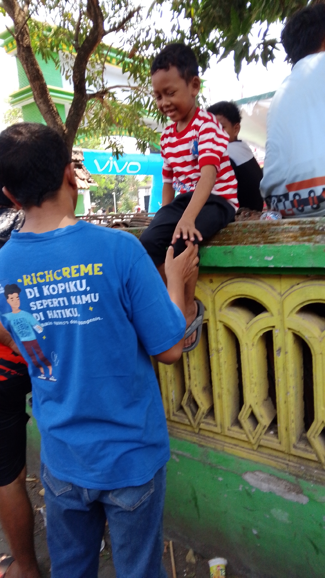 In Mancar, Indonesia, on a sunny day, two young boys are seen outdoors. One boy, wearing a red and white striped t-shirt and black pants, sits on a decorative yellow wall with green trim. He is looking down and smiling, with his hands resting on his knees. Behind him, another boy, partially visible, is wearing a black and white t-shirt. To the left, a third boy, wearing a blue t-shirt with yellow text and graphics, and blue jeans, stands facing away from the camera. The blue t-shirt has text that appears to be an advertisement. The background features trees, a building with a "VIVO" banner, and more people. The ground is covered in dirt and debris.