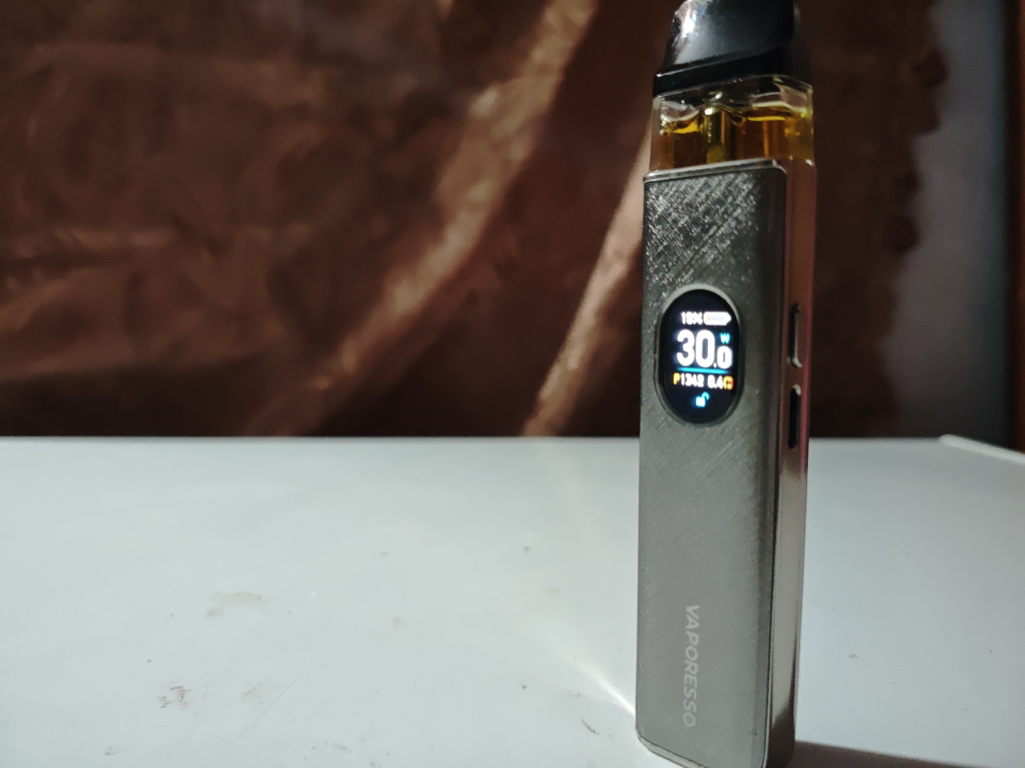 This is a close-up, eye-level shot of a silver Vaporesso vape device sitting on a white, slightly dusty surface. The background is a blurred, textured wall in shades of brown and tan, possibly fabric or wood. The vape device is upright, and its digital screen is illuminated, displaying "18% battery", "30.0W", and "P1342 0.4Ω". The brand name "VAPORESSO" is visible vertically on the side of the device. The device has a black mouthpiece and a clear cartridge containing a yellowish liquid. The lighting appears to be artificial, casting a slight glow on the vape device and its screen. The overall scene is still and focused on the product. No people are visible. The setting appears to be indoors.