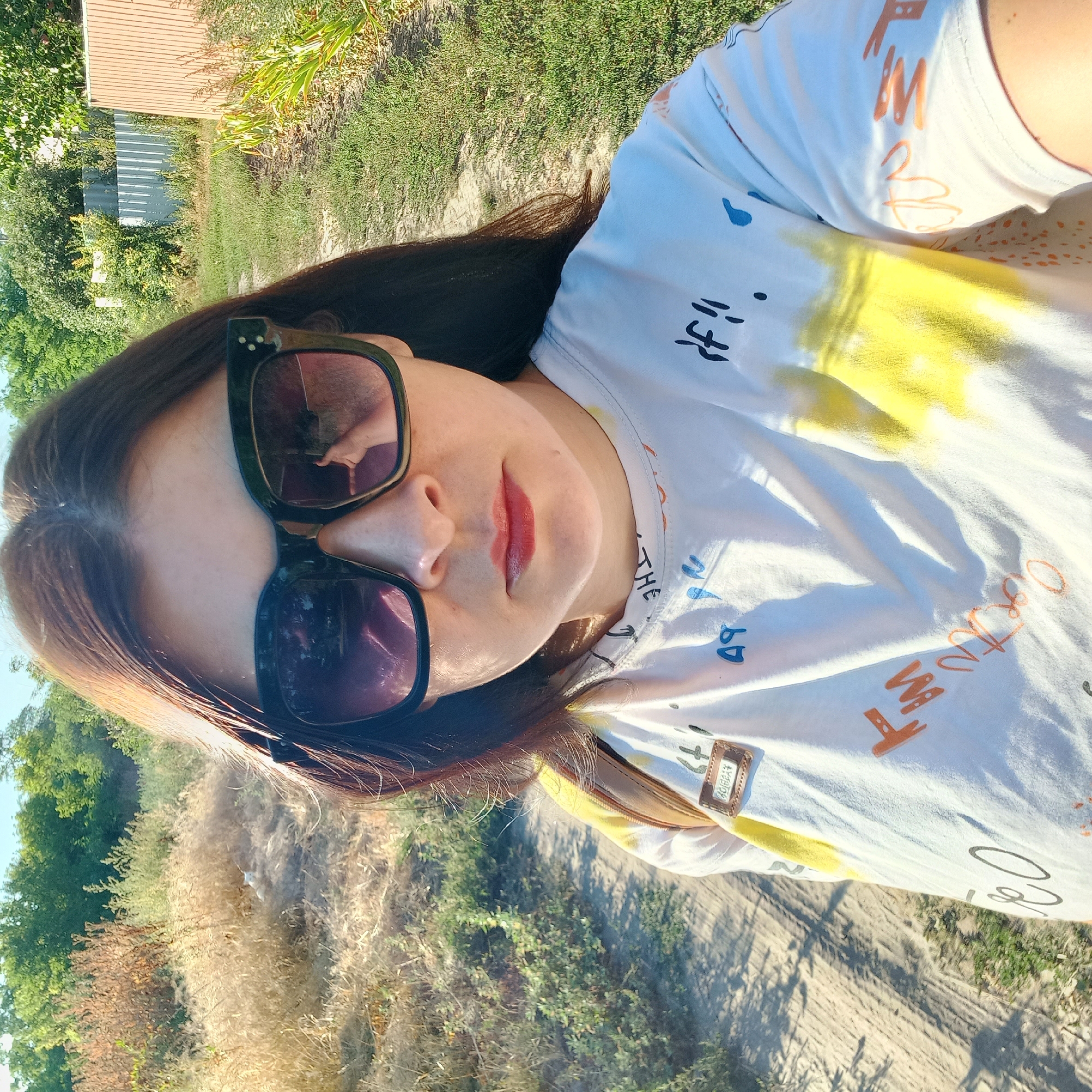 A woman with brown hair and sunglasses is taking a selfie outdoors. She is wearing a white t-shirt with colorful abstract designs. The background shows a dirt path, dry grass, green bushes, and trees. A corrugated metal fence is visible in the distance. The scene appears to be in a rural or suburban area.