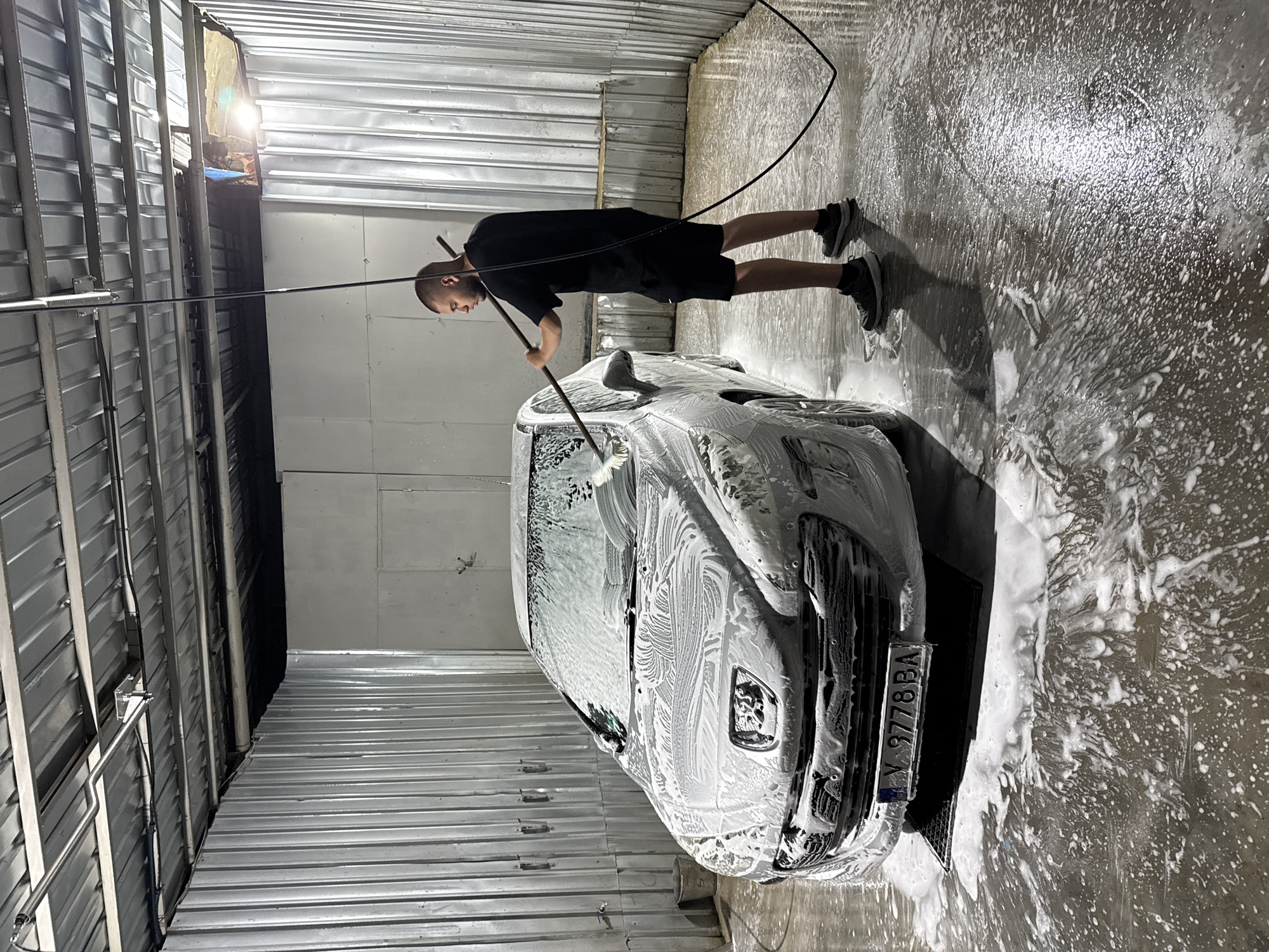 The image shows a man washing a car in a carwash with ribbed metal walls. The man, who is bald, is wearing a black t-shirt and black shorts, along with black shoes. He is holding a long-handled brush and washing the front of the silver car with soapy water. The car is almost entirely covered in white foam. The car's license plate reads "Y 9778 BA". The floor is wet with water and soap. The scene is likely in Elhovo, Bulgaria, based on the license plate information.