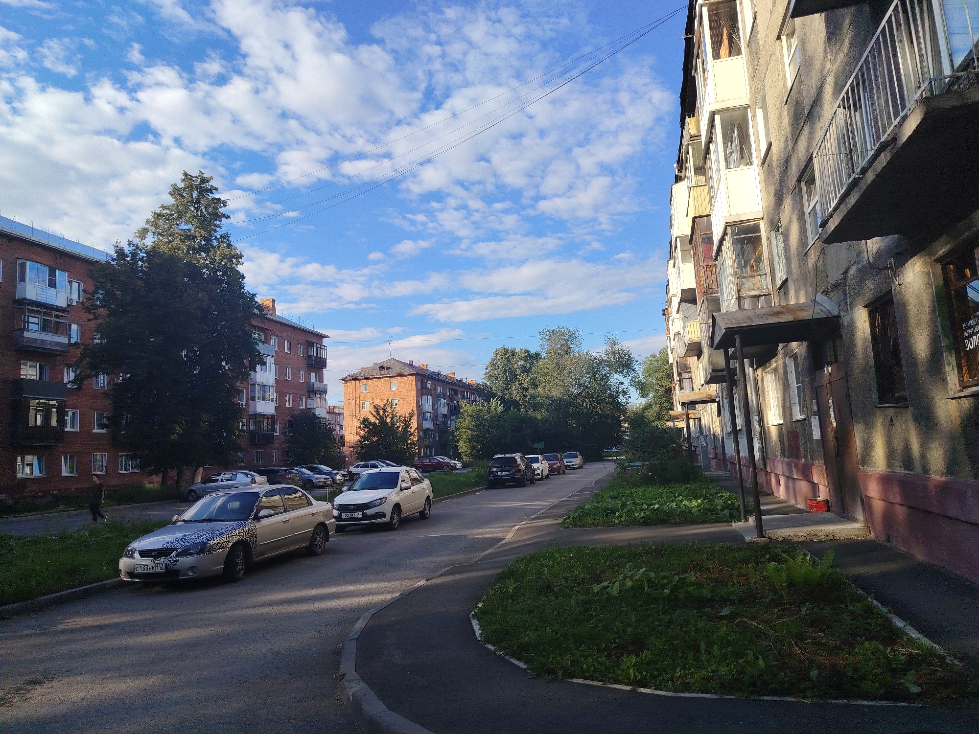 The image depicts a street scene in Kemerovo, Russia, with several apartment buildings and cars. On the left side of the image, a brick apartment building with balconies is visible. In front of it, there is a road with parked cars, including a silver sedan with a dark, patterned design on its hood. A person is walking on the sidewalk in the background.
The street curves towards the right, where another apartment building, this one with a grey facade and balconies, dominates the foreground. The building's ground floor appears to have shops or entrances, with awnings and some graffiti visible. Green grass and some plants are growing along the sidewalk and in front of the buildings.
In the background, more apartment buildings and trees are visible, with a bright blue sky filled with white, scattered clouds overhead. Power lines are strung across the sky. The lighting suggests it is daytime, likely late afternoon or early morning due to the long shadows cast by the buildings. The overall impression is that of a residential area in a Russian city.