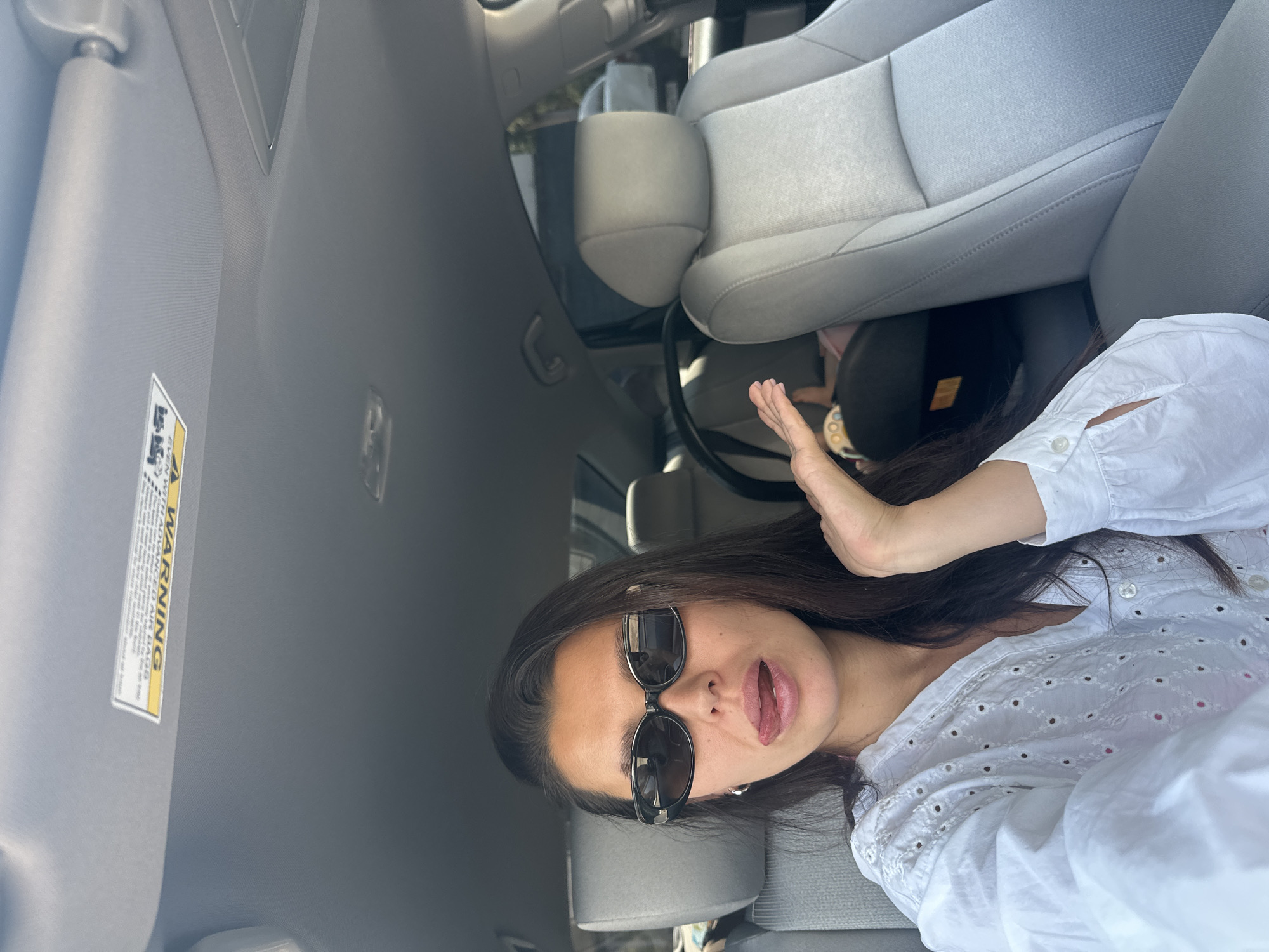 The image depicts a woman in the front passenger seat of a car, sticking her tongue out at the camera. She is wearing sunglasses and a white eyelet shirt. A child's car seat is visible in the background. The car's interior, including the dashboard and seats, is visible. The lighting suggests it is daytime. There is no information in the image to suggest the location is in Chaikovsky, Russia.