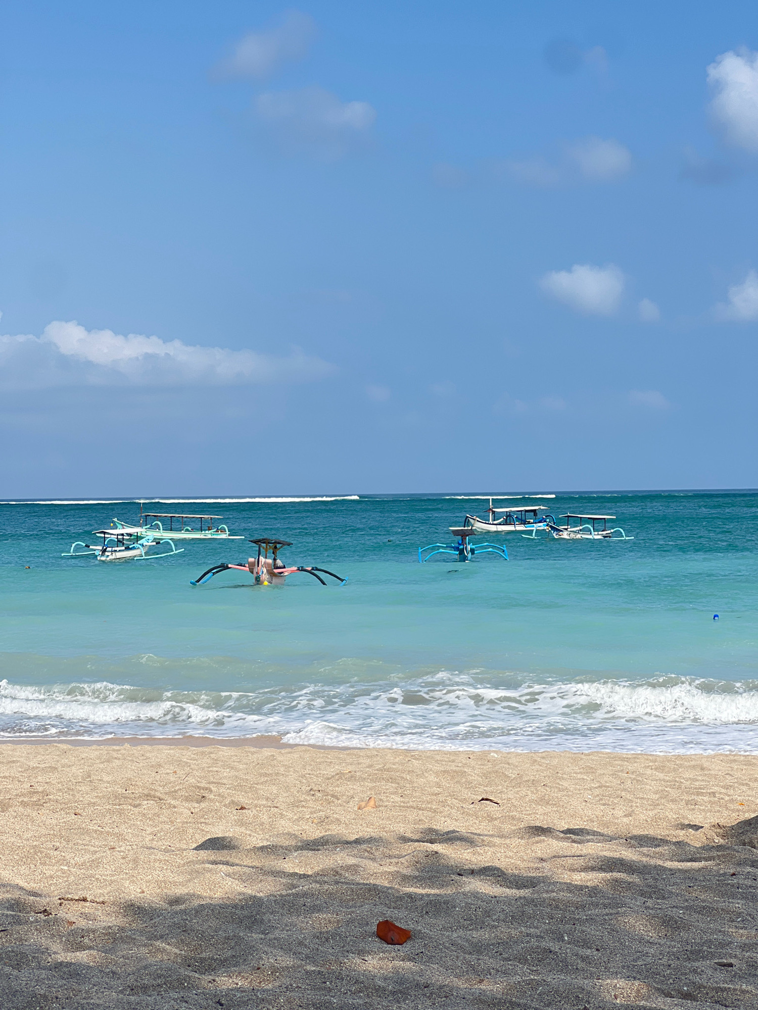 This is a full shot of a beach scene in Kuta, Indonesia. The foreground is dominated by a sandy beach with scattered dry leaves and shadows cast by an unseen object. The midground shows the ocean with gentle waves breaking and several traditional Balinese outrigger boats floating on the clear, turquoise water. The boats vary in size and color, with some having canopies. In the background, the horizon is marked by a thin white line, likely the surf break, and a bright blue sky with scattered white clouds. The scene conveys a peaceful and sunny day at the beach.