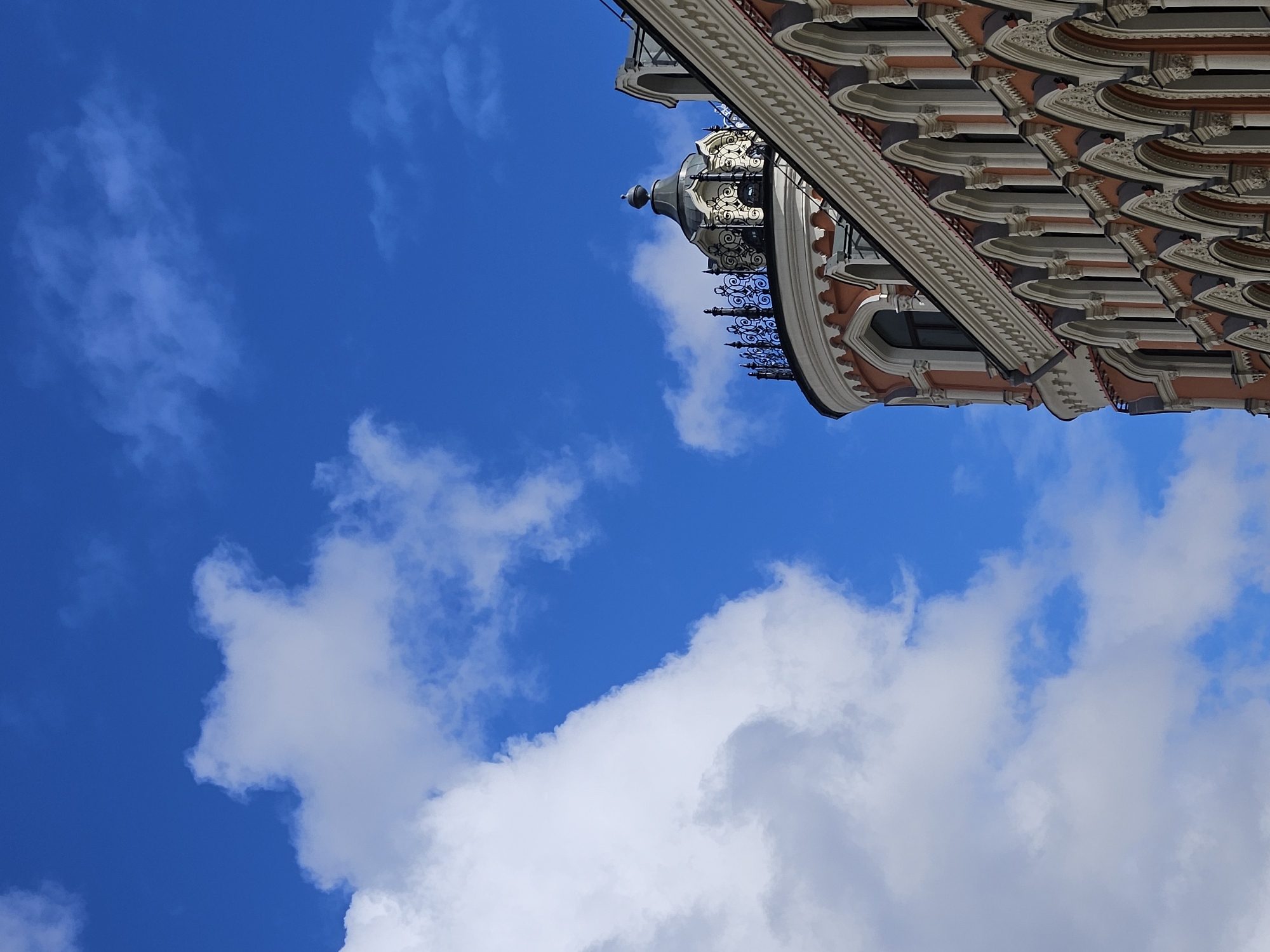 The image depicts a building exterior against a blue sky with clouds. The architectural style appears ornate and classical, with light-colored facade elements, intricate details, and a dome. The location is likely Moscow, Russia, based on the architectural style. The image is rotated, giving the impression that the building is leaning diagonally into the sky.
