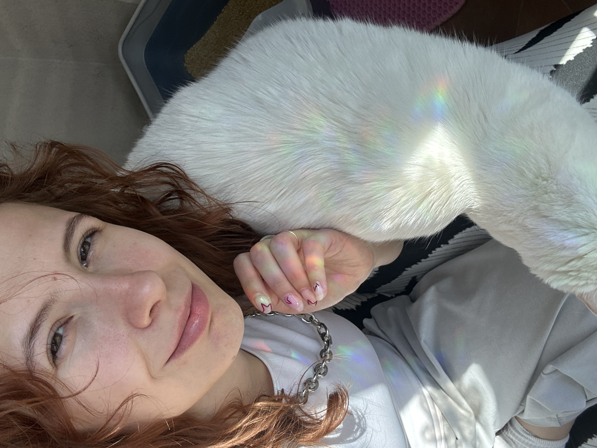 A young, light-skinned woman with reddish-brown curly hair is lying down, facing the camera. A large, white cat rests on her upper arm and torso. The woman wears a white shirt and a silver chain necklace. Her fingernails are painted with a subtle design. A light prism effect is visible on the cat's fur, the woman's hand, and shirt. Background objects include striped cushions and bedding. No information is available within the image to verify a location in Novi Sad, Serbia.