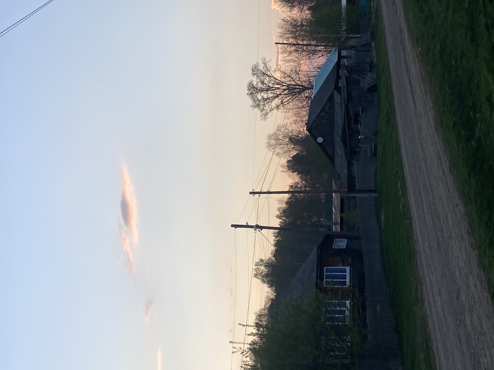 The image presents a rural scene in Узяр, Russia, at dusk. Two wooden houses are visible, weathered and dark, with window frames painted blue. An unpaved road runs alongside the houses, bordered by green grass and foliage. Utility poles with power lines stretch across the frame, silhouetted against the sky. The sky itself is a gradient of light blue to orange, with wisps of cloud. A satellite dish is mounted on one of the houses. Trees with bare branches are interspersed with the houses.
