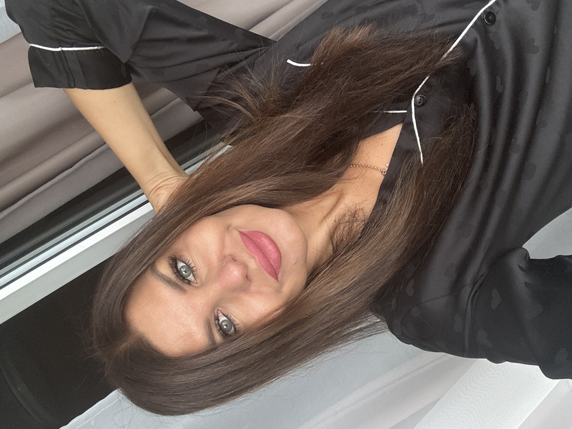 The image is a selfie of a woman with long brown hair and blue eyes, wearing black pajamas with white trim and a heart pattern. She is smiling and has her arm behind her head. The background consists of curtains and a window. The scene is indoors.