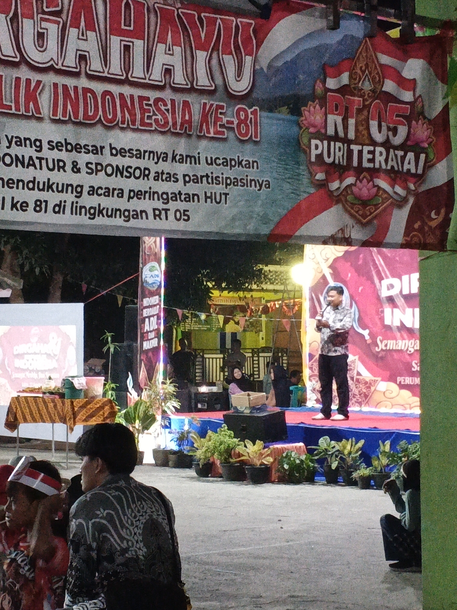 The image captures an outdoor event in Cikande, Indonesia, likely a celebration of Indonesian Independence Day. A large banner at the top reads "MEGAHAYU" and "LIK INDONESIA KE-81", indicating the 81st anniversary of Indonesian Independence. Below this, text acknowledges donors and sponsors for their participation in the event commemorating HUT (Hari Ulang Tahun - Birthday) ke-81 in the RT 05 (Rukun Tetangga - Neighborhood Association 05) environment.
In the center of the scene, a man stands on a stage, holding a microphone and speaking. He is wearing a patterned shirt and dark pants, with a small bag slung across his chest. The stage is decorated with plants in pots and a large pink backdrop featuring text that appears to say "DIF INI Semanga S: PERUM". To the left of the stage, a table is set up with food or offerings, covered by a patterned cloth. Another banner, vertically oriented, hangs to the left of the stage, with text including "INDONESIA BERDAULAT ADIL DAN MAKMUR".
In the foreground, several people are visible. Two individuals, one wearing a patterned shirt and the other with a headband featuring the Indonesian flag colors, are in the lower left. Another person is seated on the ground in the lower right corner. The environment suggests a community gathering, with multiple stages, banners, and seating areas. The lighting suggests it might be late afternoon or early evening.