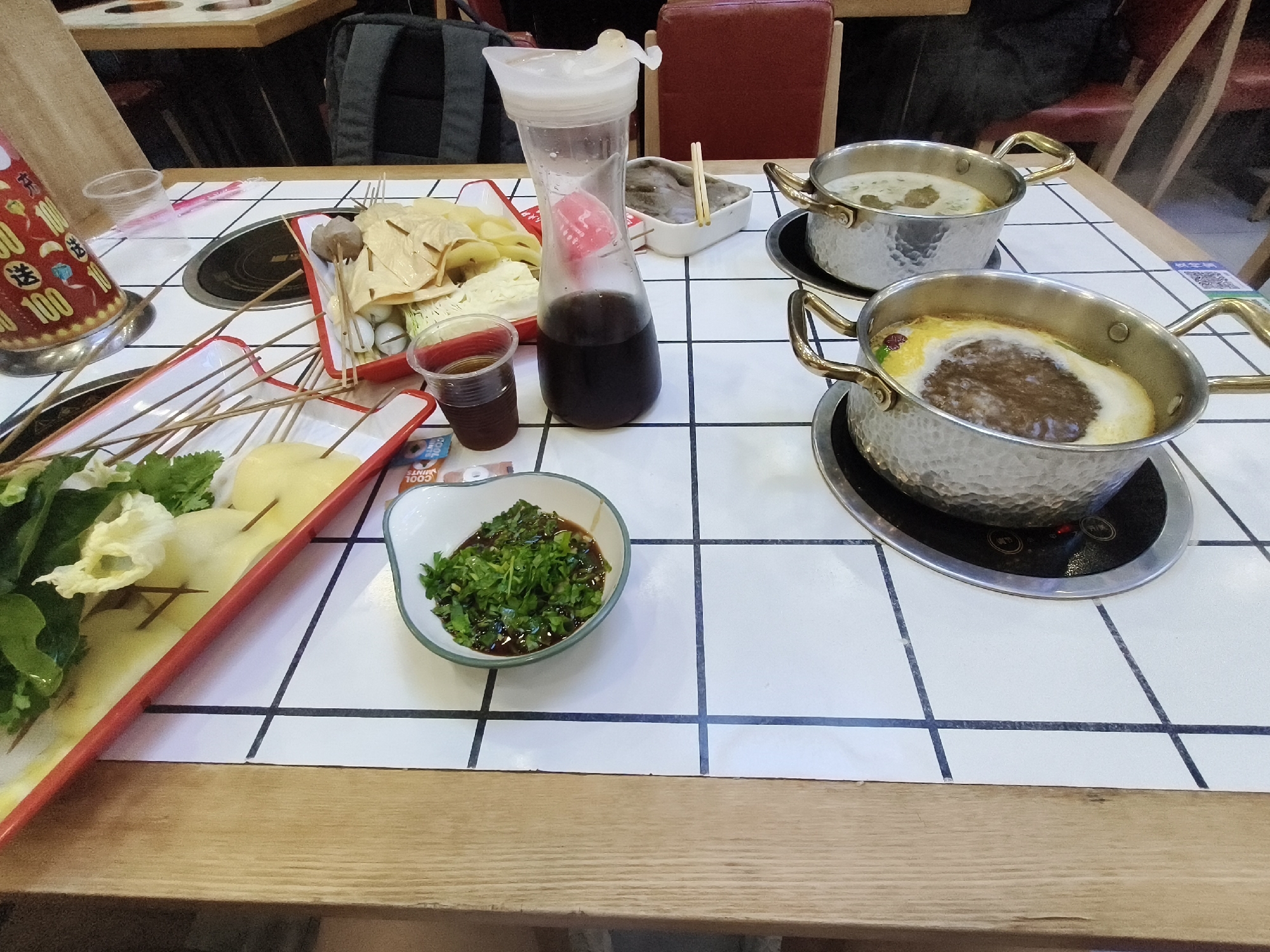 The image depicts a tabletop setting for a meal, likely at a restaurant. The table is covered with a white tiled surface with black grid lines. On the table, there are two metallic pots with handles, containing soup or broth, and various food items served on red and white trays. These food items include what appear to be skewers with meatballs, tofu, and vegetables, as well as some leafy greens. A clear plastic cup with a dark liquid is also visible, along with a tall, clear pitcher filled with a dark liquid and a pink object. A small white bowl contains chopped green herbs. In the background, parts of chairs and a backpack can be seen, suggesting an indoor dining environment. The overall scene is a close-up view of a meal being prepared or served, with a focus on the food and dining accessories. The image does not contain specific landmarks or elements that would allow for precise identification of its location within Mudanjiang, China.