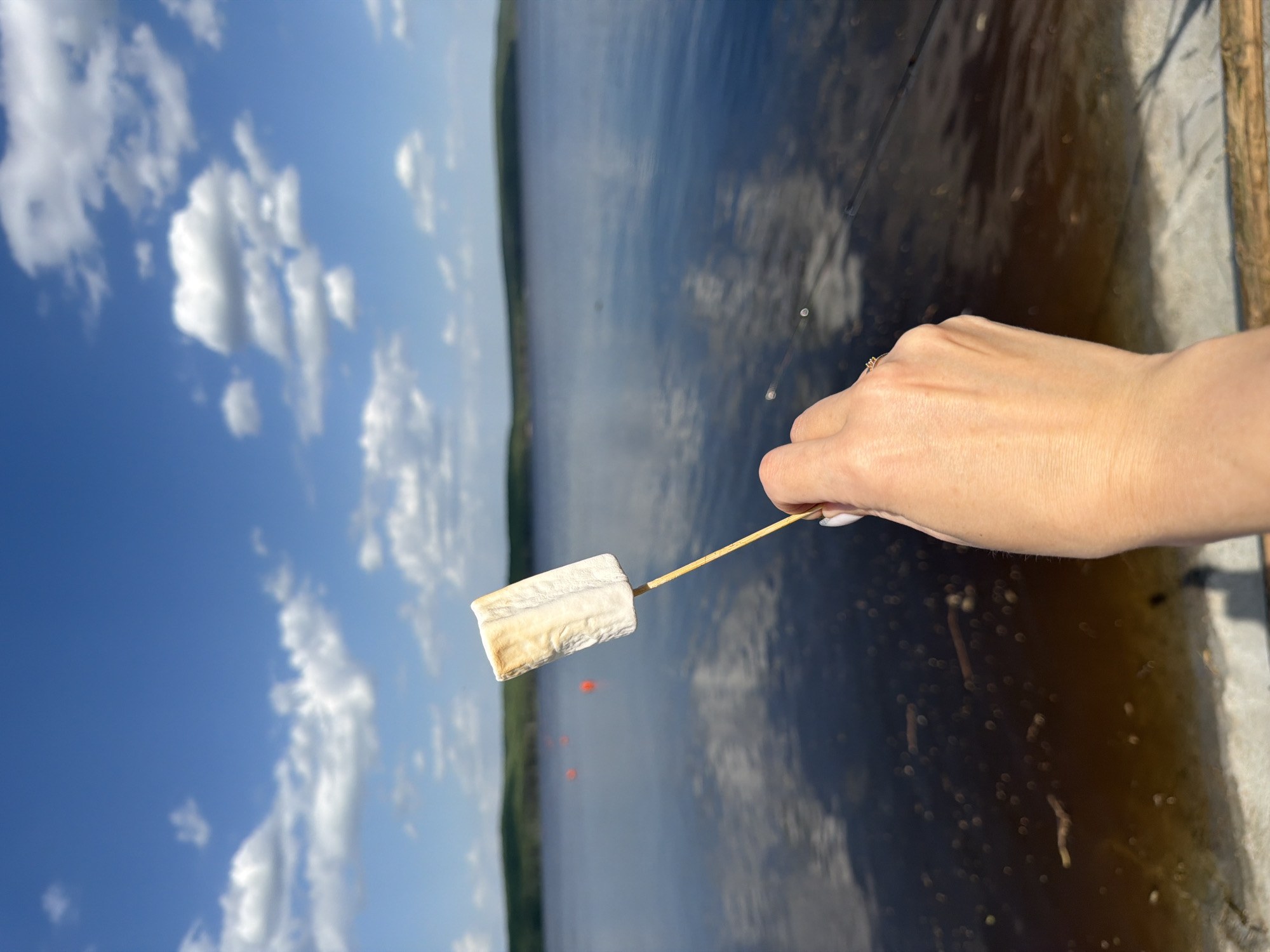 A person holds a roasted marshmallow on a wooden skewer over the edge of a concrete platform. Behind them is a body of water, possibly a lake or reservoir, with a visible shoreline in the distance. The sky is blue with scattered white clouds. A cluster of orange buoys is visible in the water. The location may be near Chaikovsky, Russia, considering the terrain and geographical features.