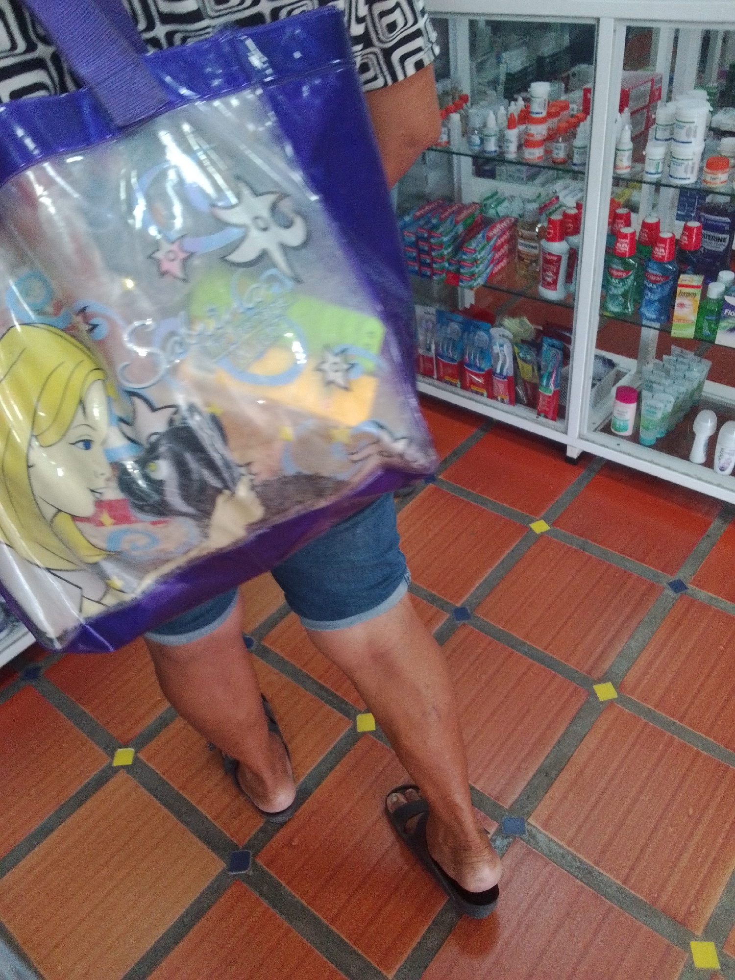 In Juan Griego, Venezuela, a person stands on a tiled floor in front of a display cabinet filled with health and beauty products. The person is holding a transparent blue tote bag with a cartoon image of a blonde woman. They are wearing denim shorts and flip-flops. The display cabinet contains various items, including toothbrushes, toothpaste, mouthwash, and medications. The floor is made of terracotta-colored tiles with a pattern of blue and yellow accents.