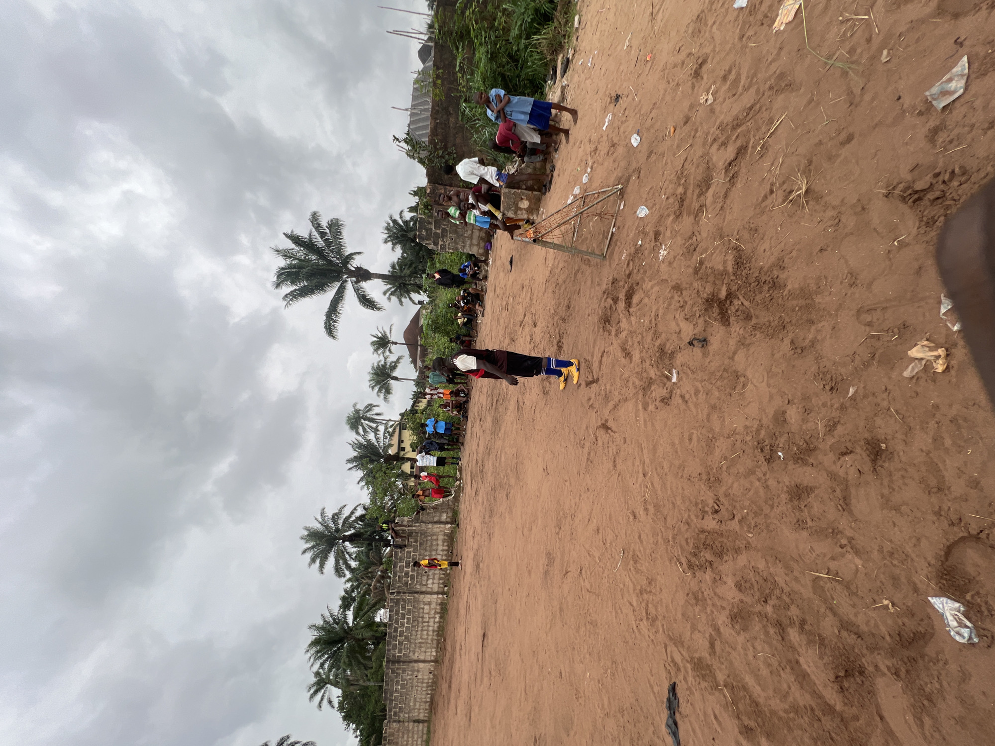 The image depicts a soccer match in progress on a sandy field in Nkpor, Nigeria. The field is lined with palm trees and a brick wall. A group of people are gathered near one of the goals. Litter is scattered across the field. Overcast sky.